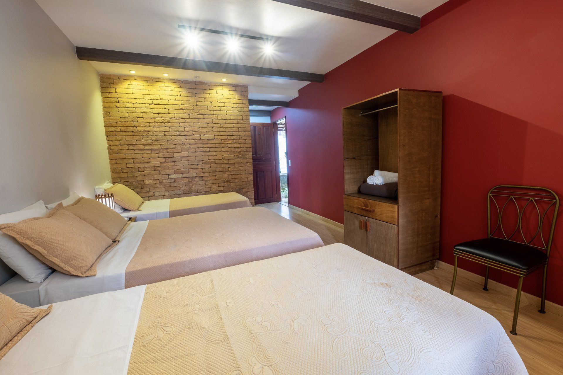 Quarto com três camas, paredes em tons de vermelho e bege, detalhes em tijolo aparente, guarda-roupa de madeira e uma cadeira decorativa.