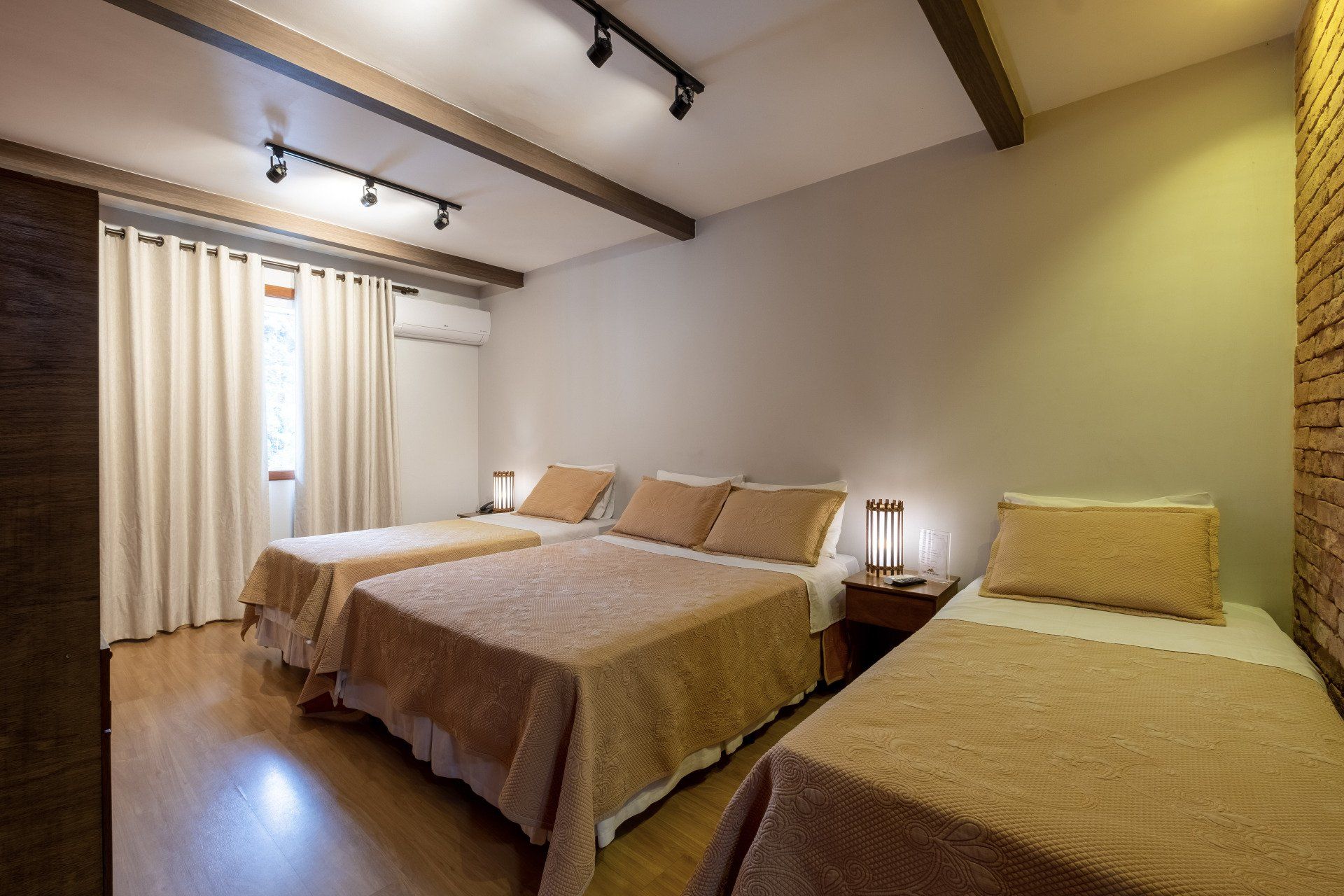 Quarto com três camas, roupa de cama bege, vigas de madeira e parede de tijolos.