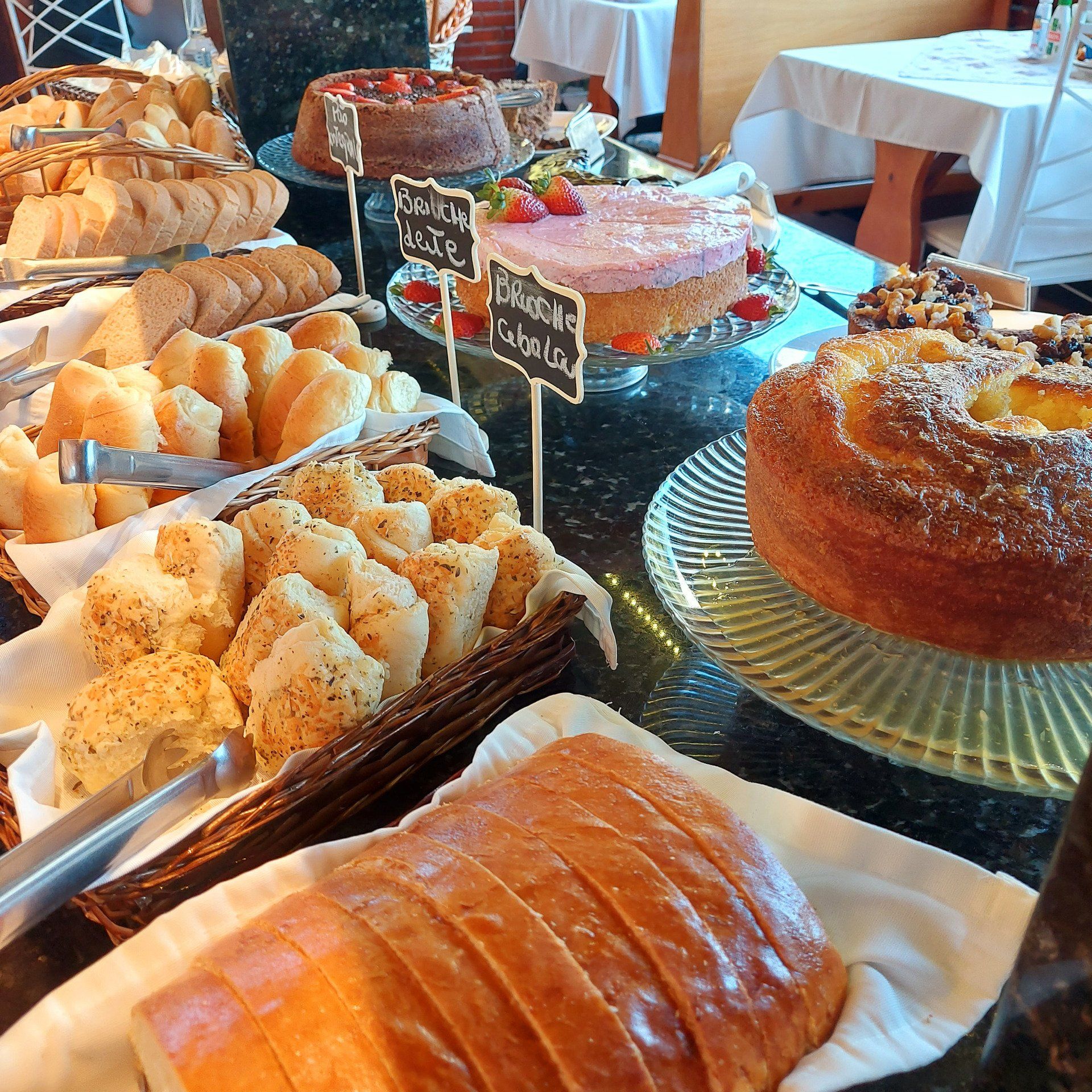 Buffet de café da manhã com diversos tipos de pães, bolos e doces em uma bancada preta.