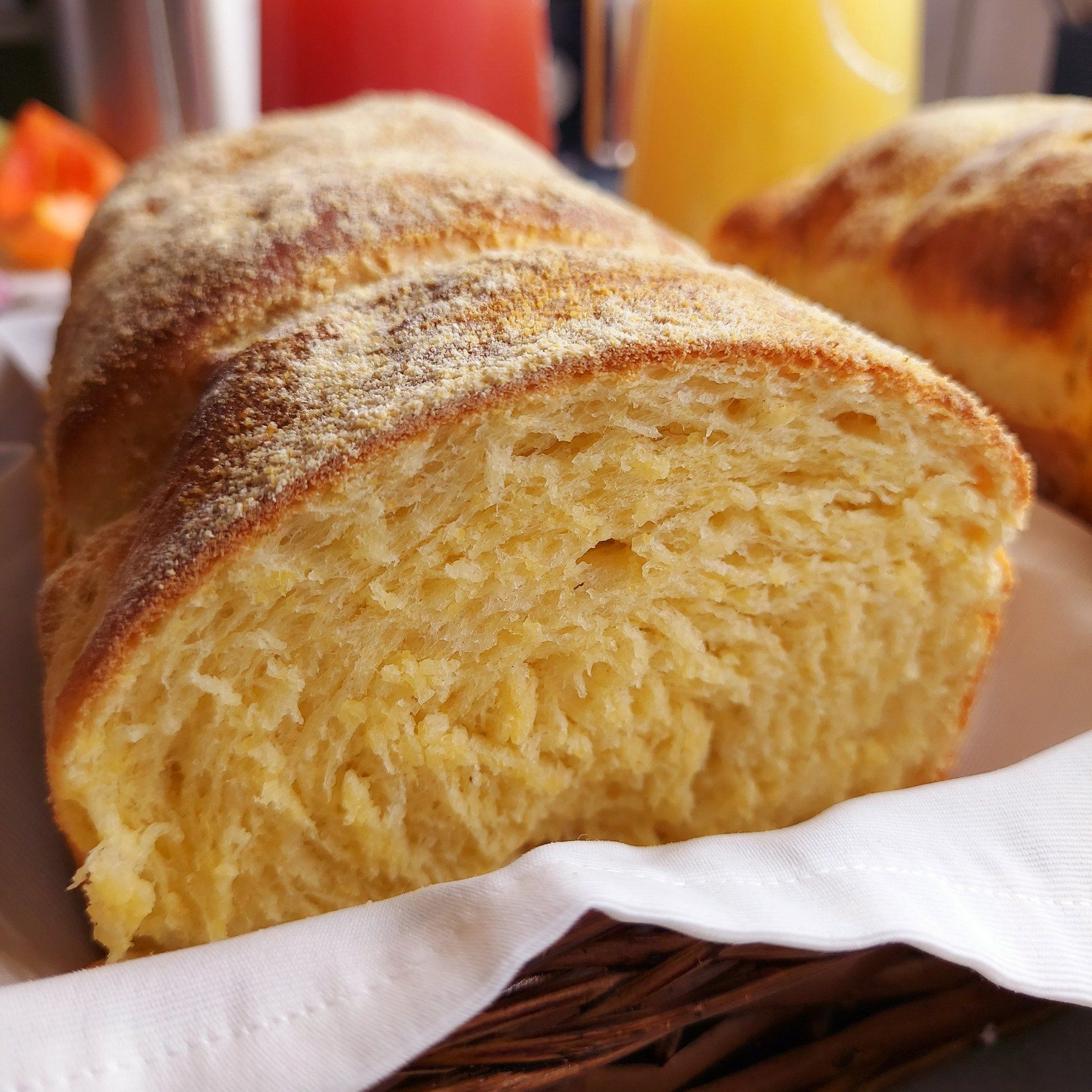Pão amarelo-dourado, fatiado para mostrar a textura macia, em uma cesta com guardanapo; suco ao fundo.