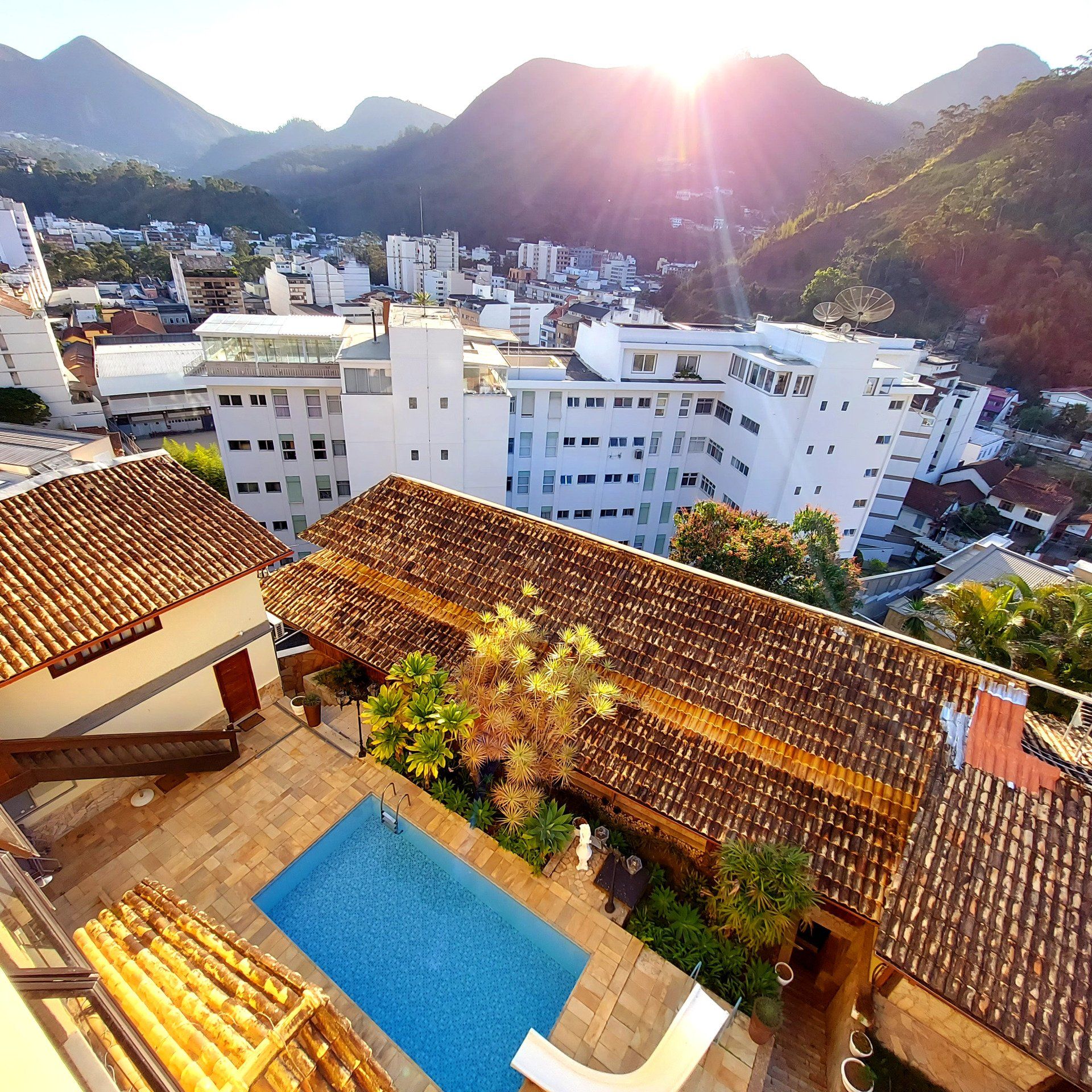 Paisagem urbana com piscina e montanhas. Prédios, piscina, telhados marrons, árvores, luz solar.