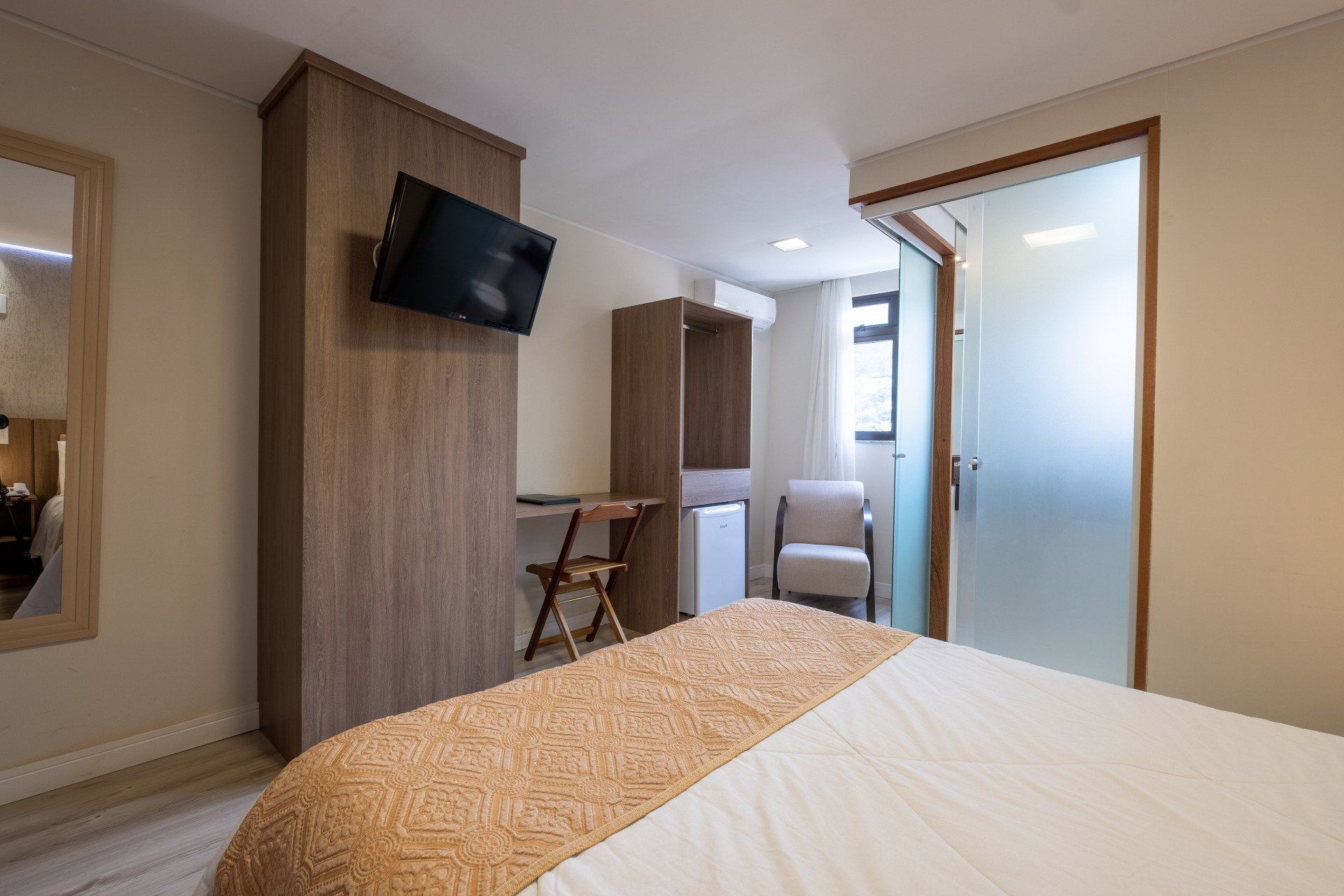 Quarto de hotel com cama, guarda-roupa, escrivaninha, cadeira, TV e porta de vidro fosco.