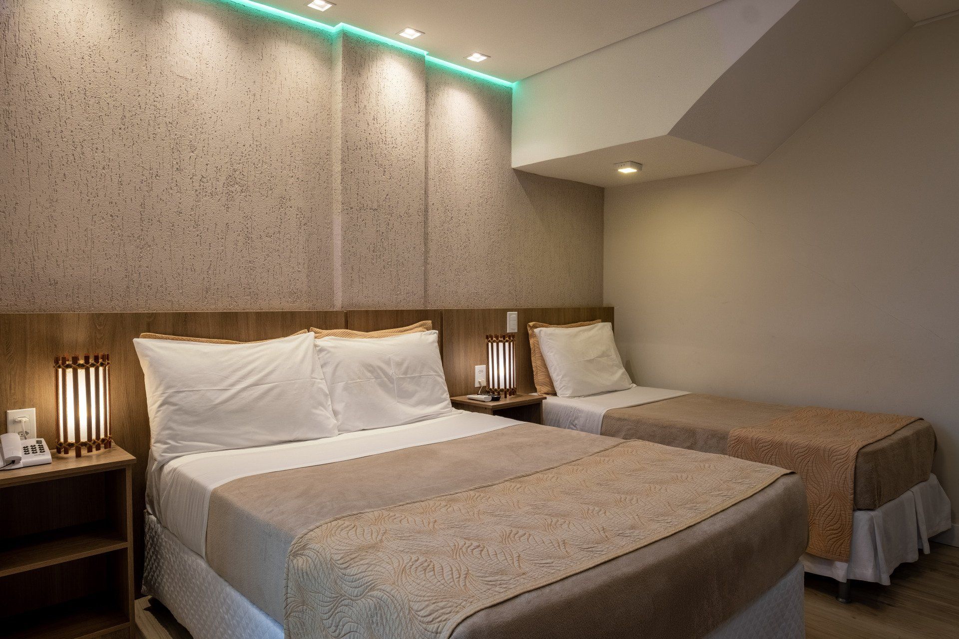 Quarto de hotel com duas camas, tons neutros, cabeceira de madeira, abajures e iluminação decorativa.