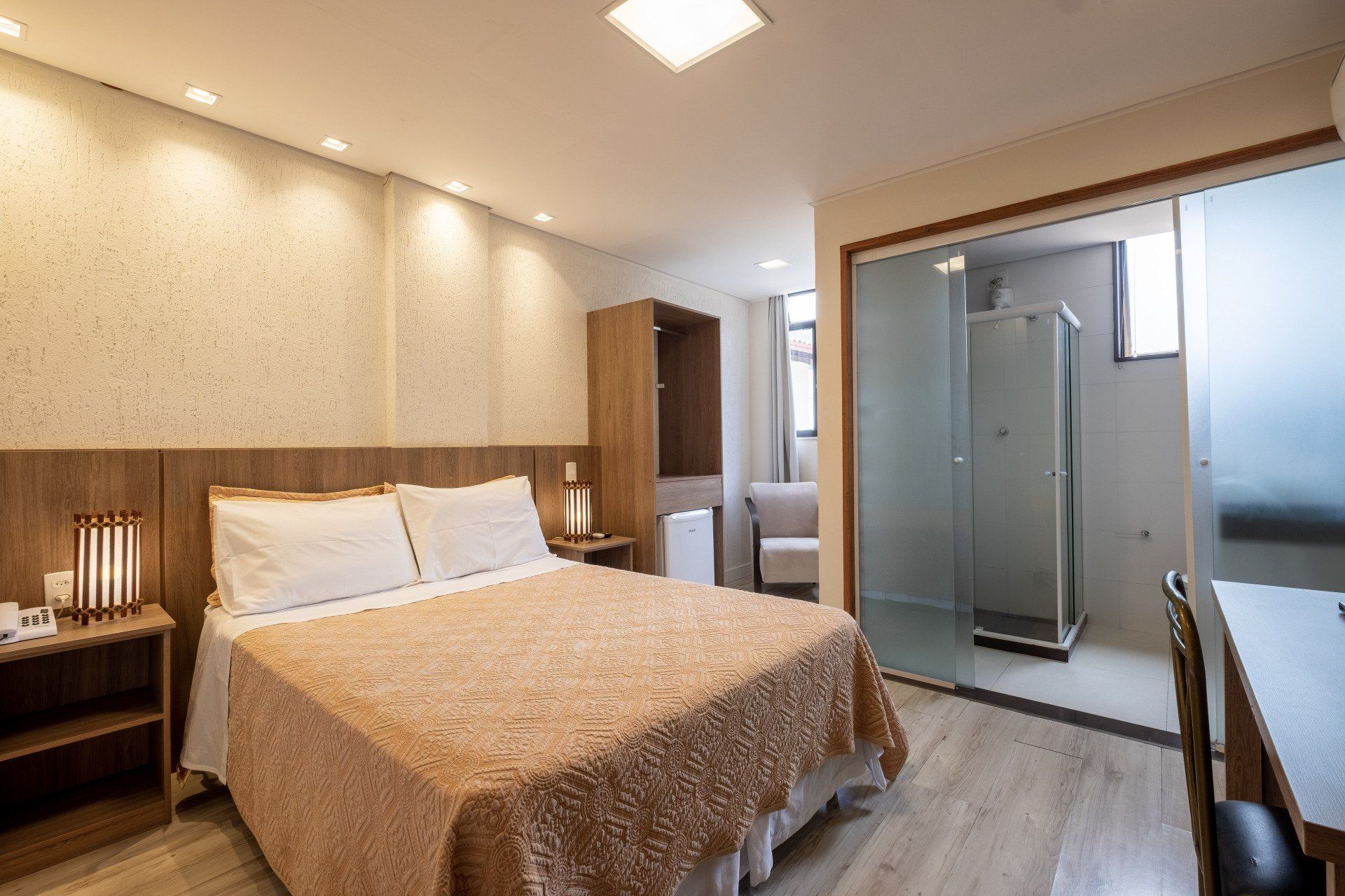 Quarto de hotel com cama, cabeceira de madeira e porta de vidro deslizante para o banheiro.