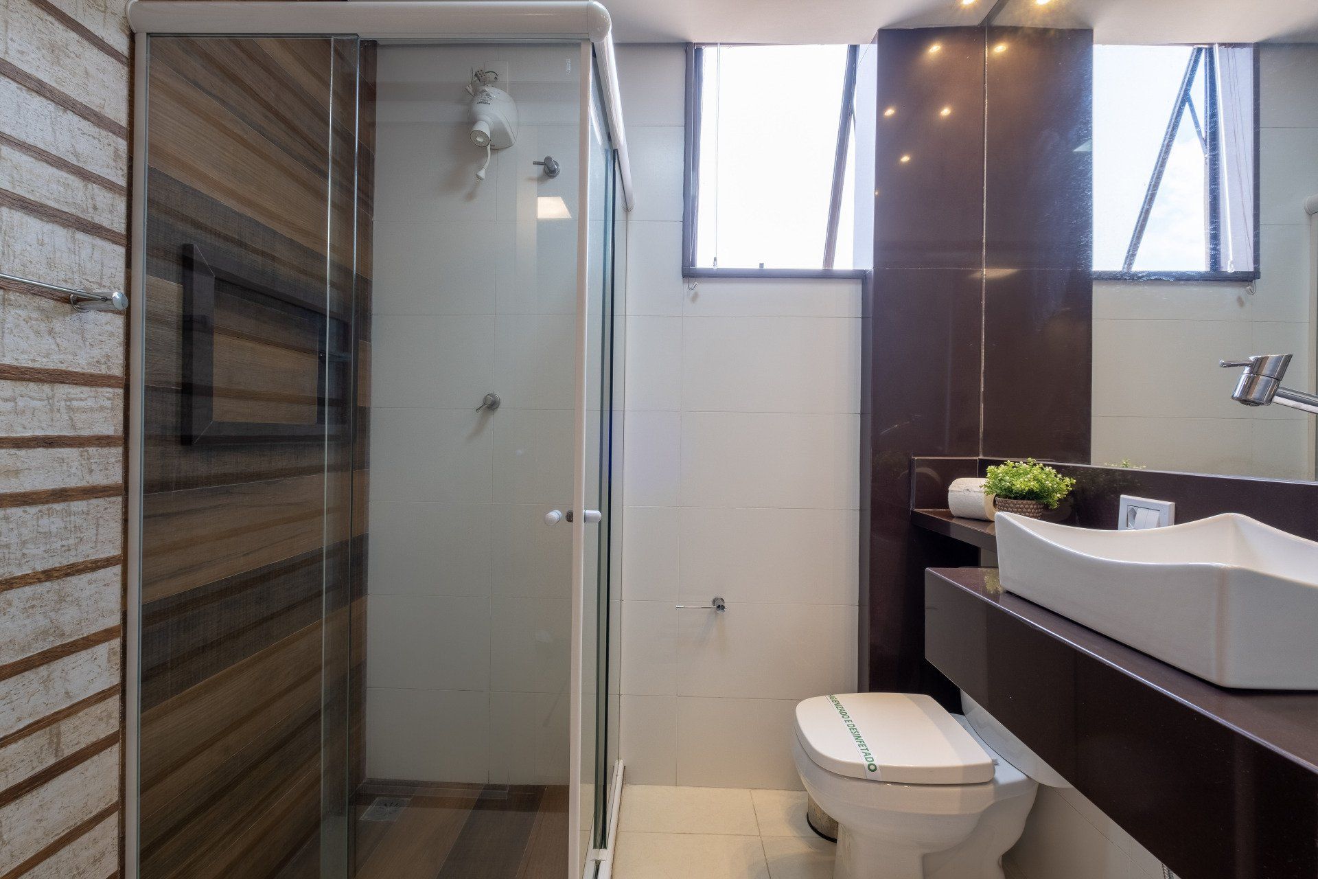 Banheiro moderno com box de vidro, vaso sanitário e pia de sobrepor. Parede e janela com detalhes em marrom.
