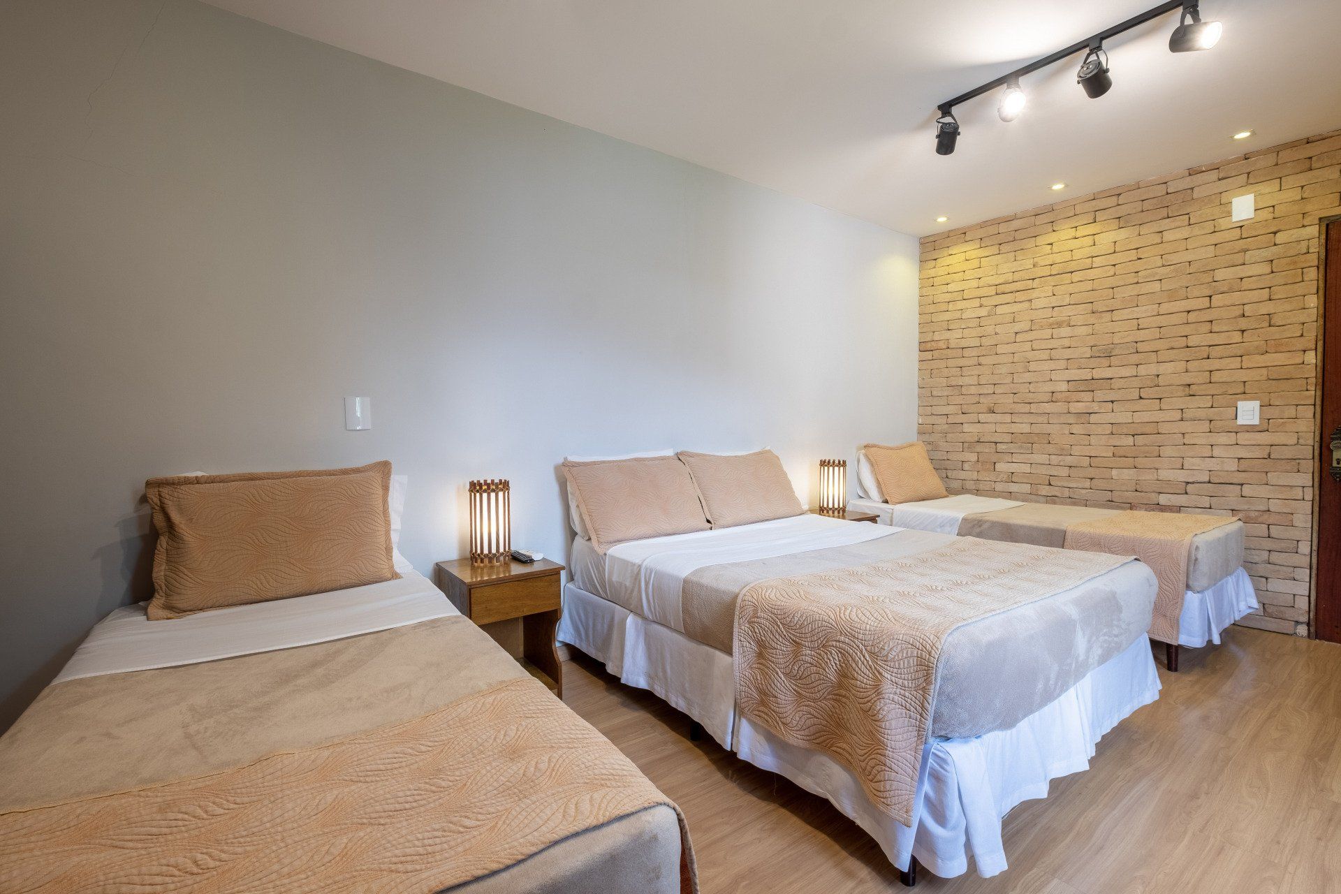Quarto com três camas: uma de solteiro, uma de casal e outra de solteiro; lençóis bege, parede de tijolos à vista.