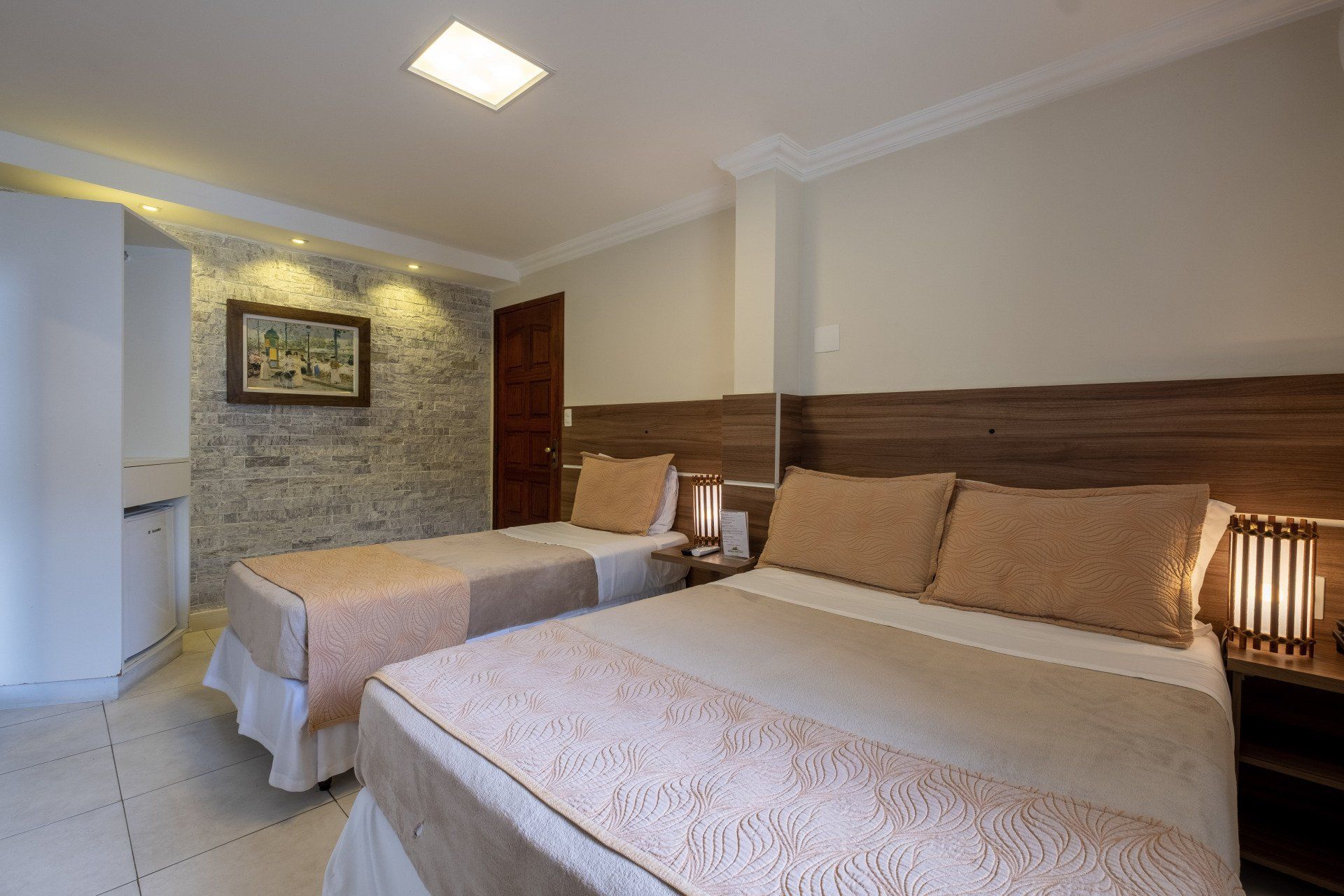 Quarto de hotel com duas camas, uma de casal e uma de solteiro, paredes em tons neutros, detalhes em pedra, obras de arte e iluminação aconchegante.