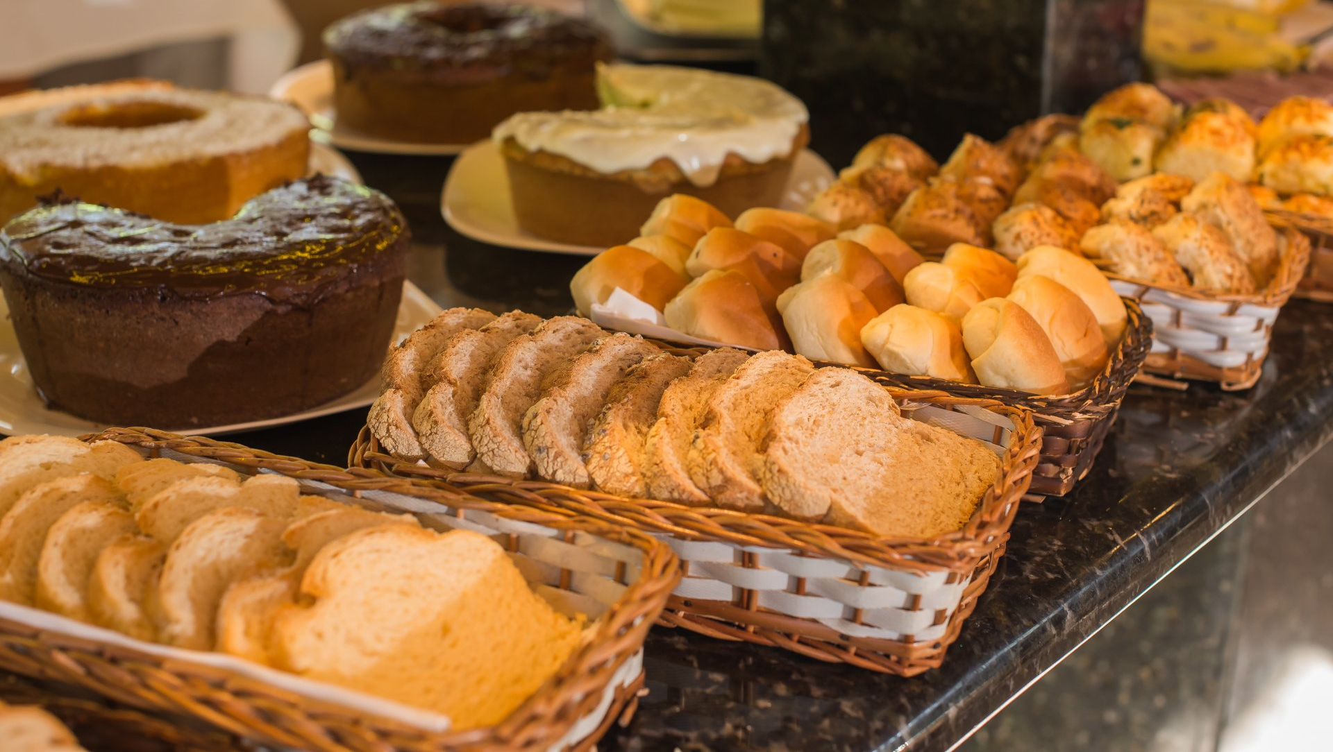 Buffet de café da manhã com diversos pães, bolos e pãezinhos em cestas de vime sobre uma bancada escura.