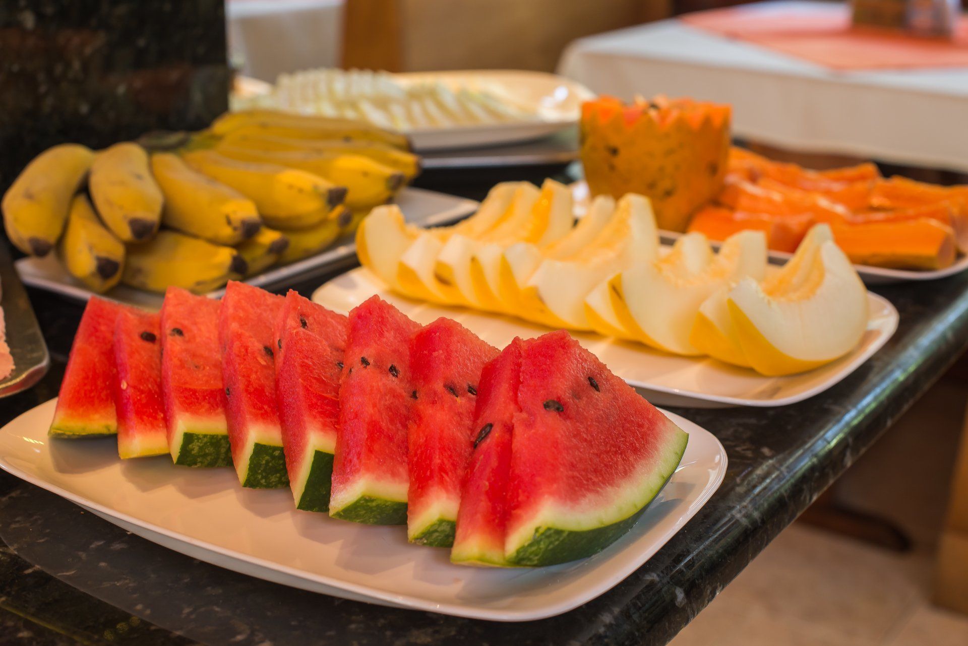 Bandejas de frutas fatiadas, incluindo melancia, banana e melão, em um bufê.
