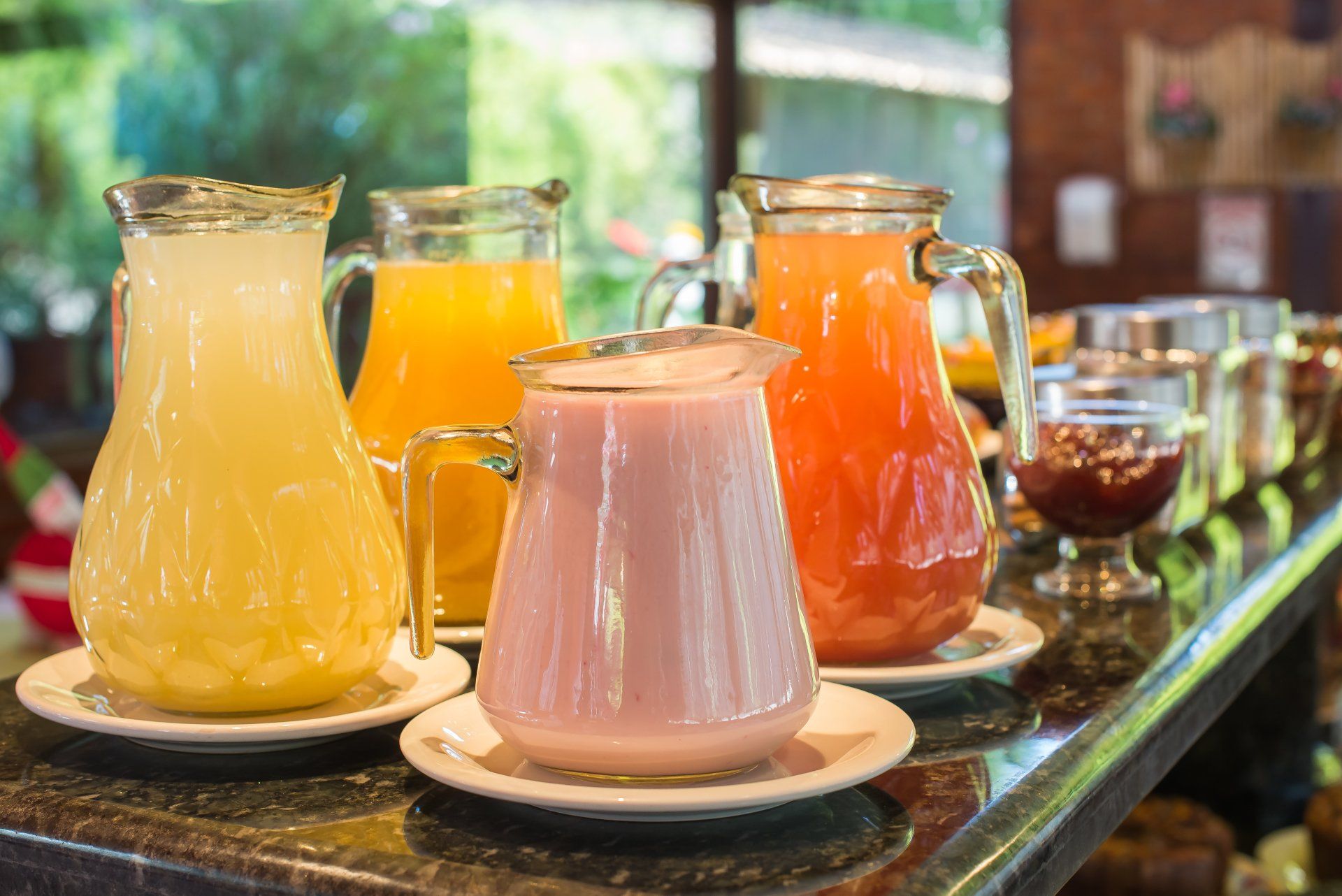 Quatro jarras de vidro com suco, uma amarela, uma laranja e uma rosa, sobre um balcão.
