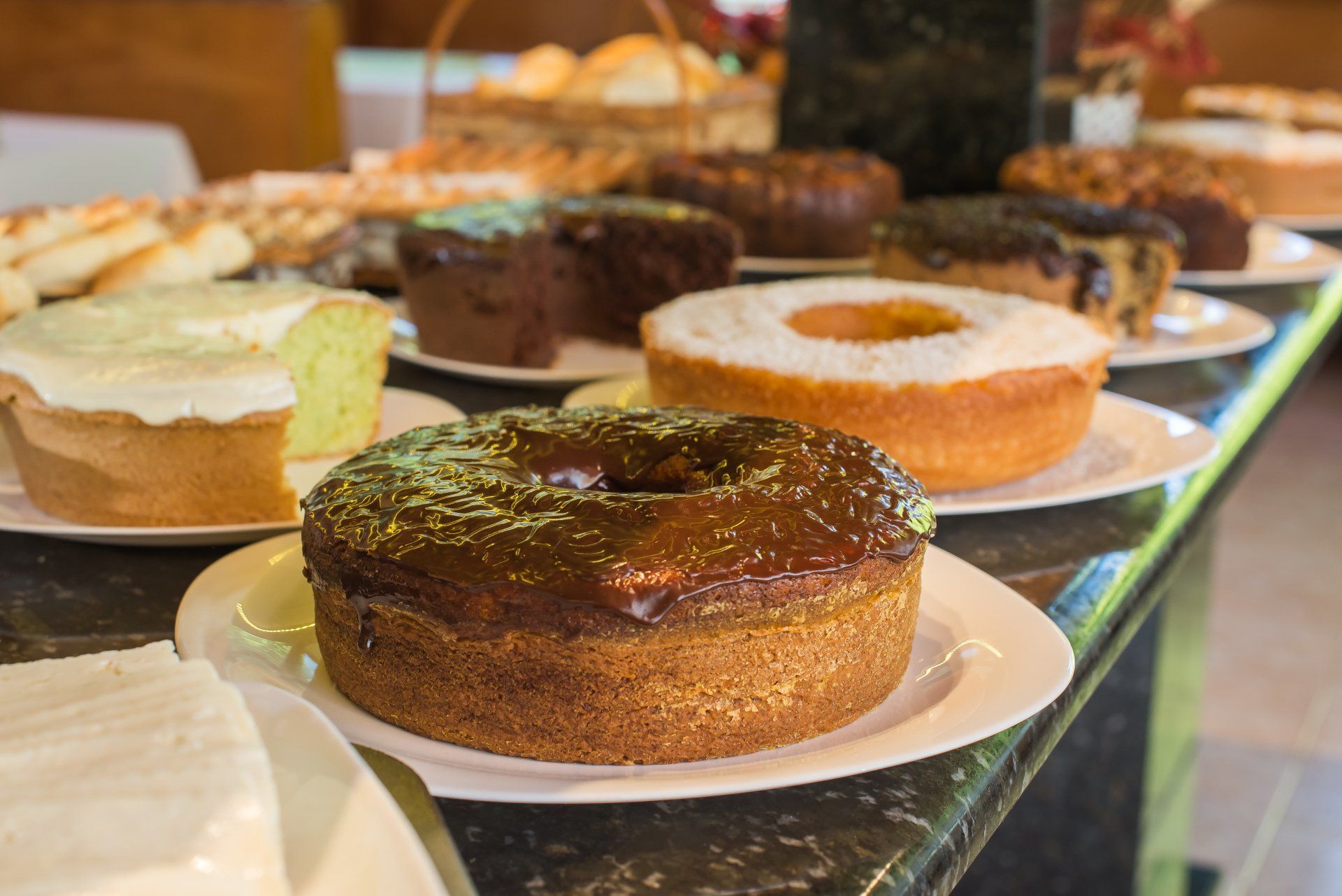 Bolos em exposição, de diversos sabores e formatos, dispostos em pratos brancos sobre um balcão.