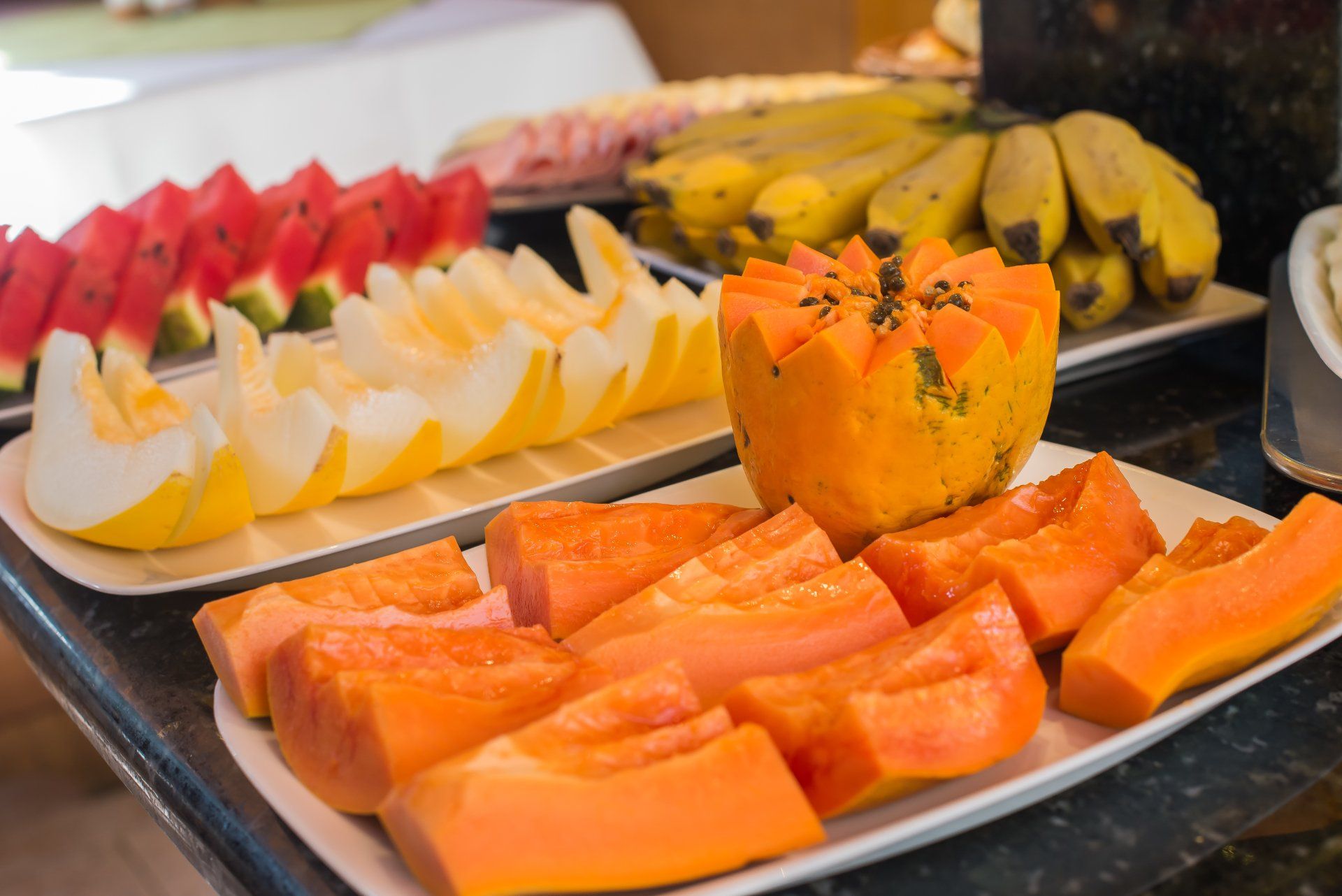 Bandejas de frutas frescas, incluindo melancia, melão, bananas e mamão, dispostas em um bufê.