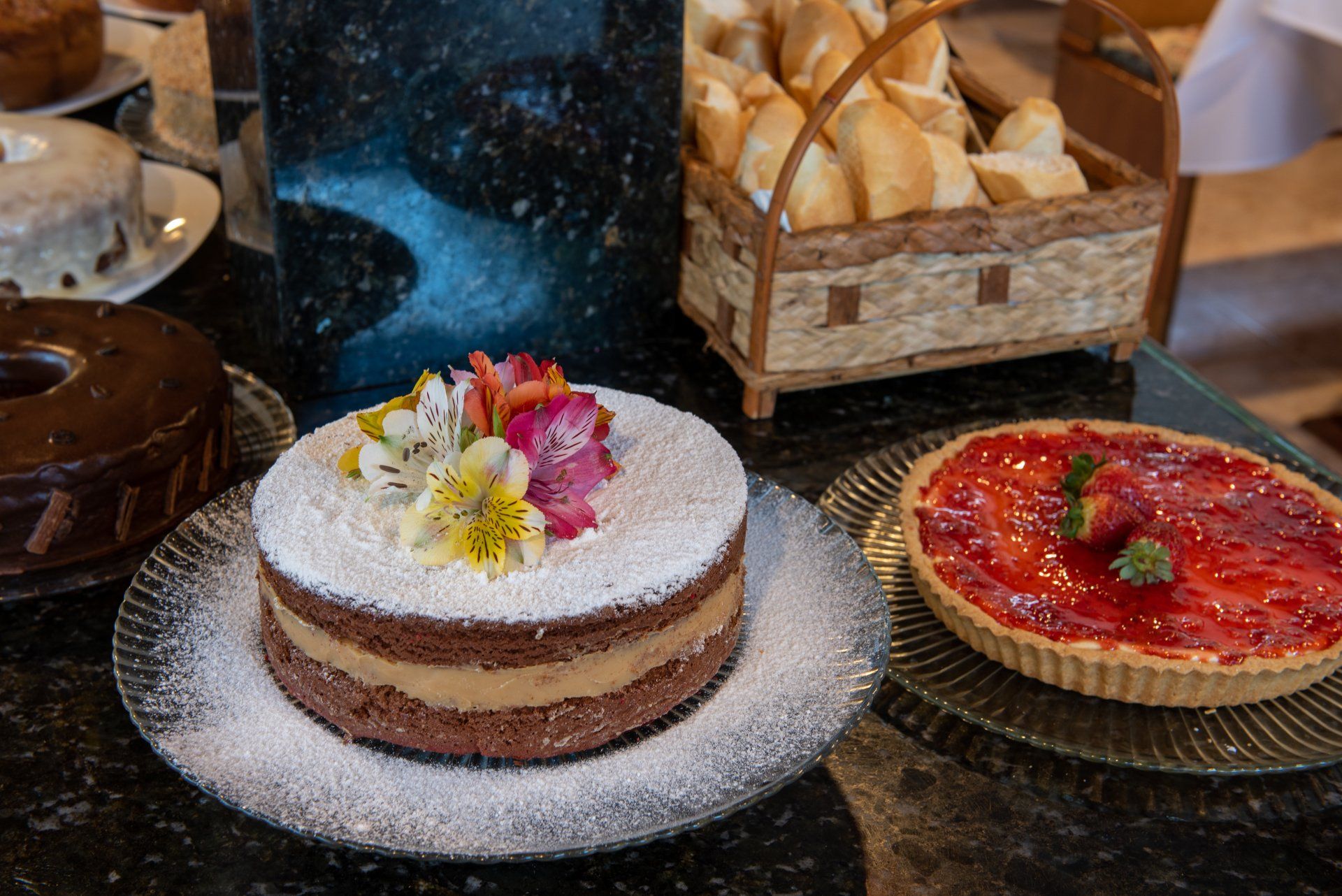 Uma vitrine de sobremesas: bolo de chocolate, bolo em camadas com flores e torta de morango; uma cesta de pães.