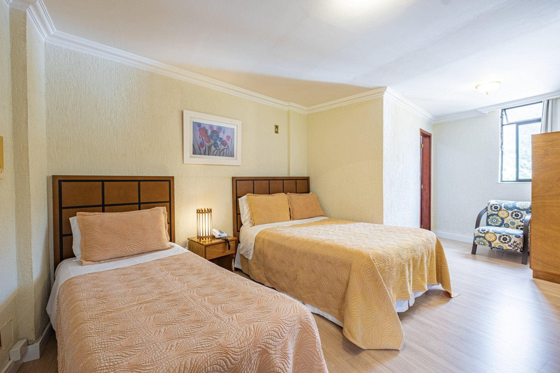 Quarto com duas camas, roupa de cama bege, uma mesa de cabeceira e uma cadeira estampada perto da janela.