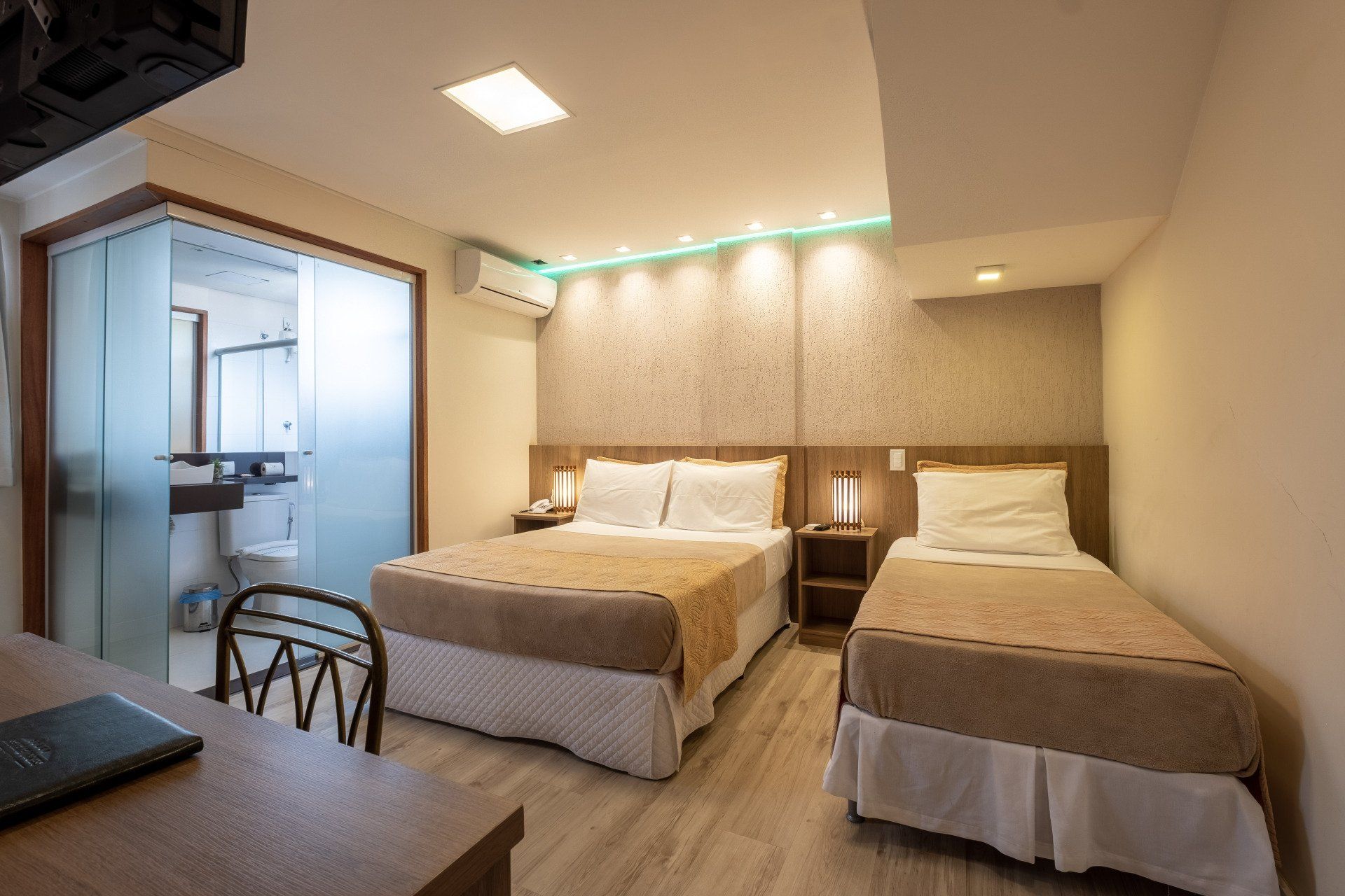 Quarto de hotel com uma cama de casal e uma de solteiro, banheiro com porta de vidro fosco e escrivaninha.