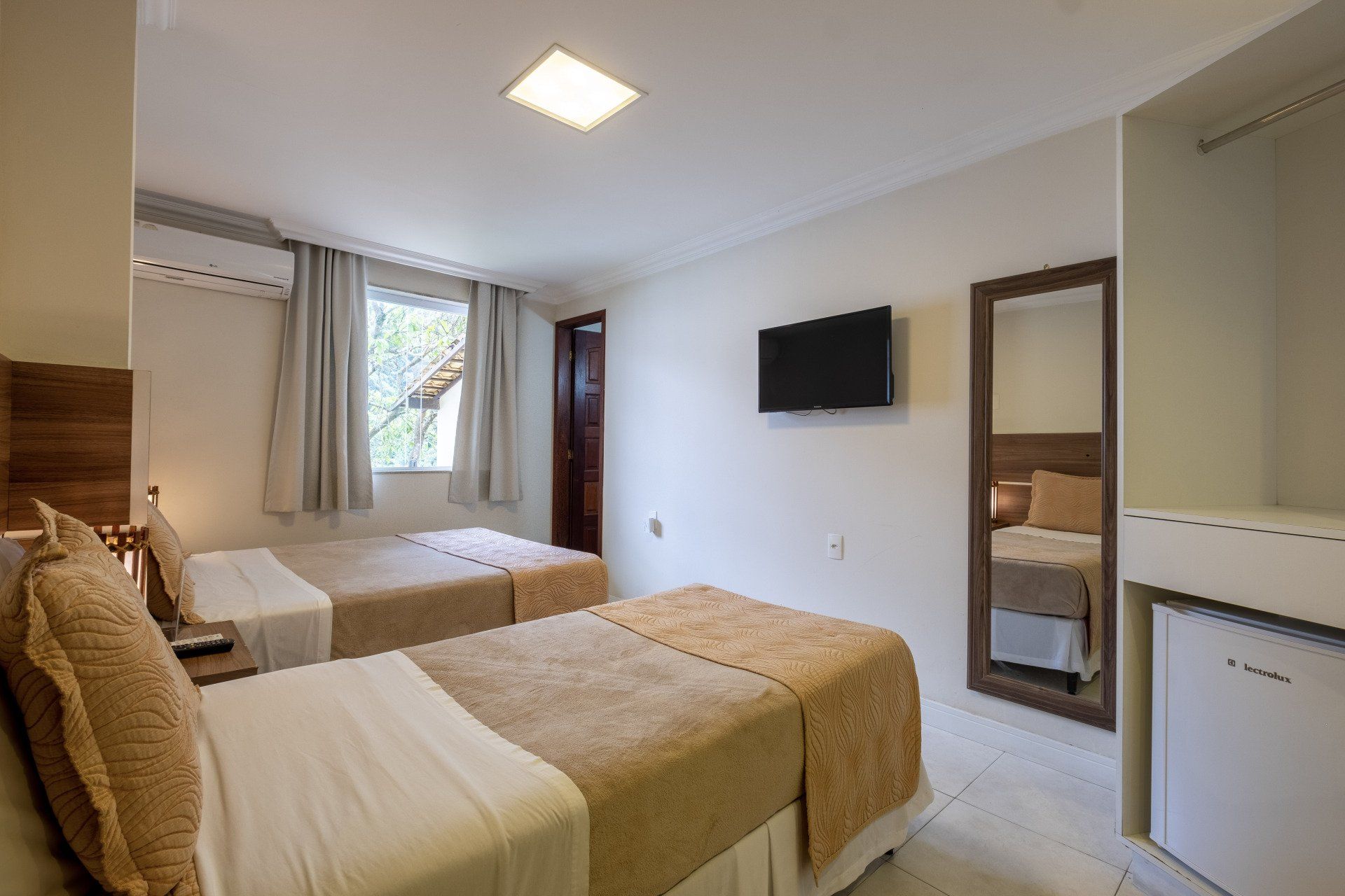 Quarto de hotel com duas camas de solteiro, TV, espelho e frigobar.