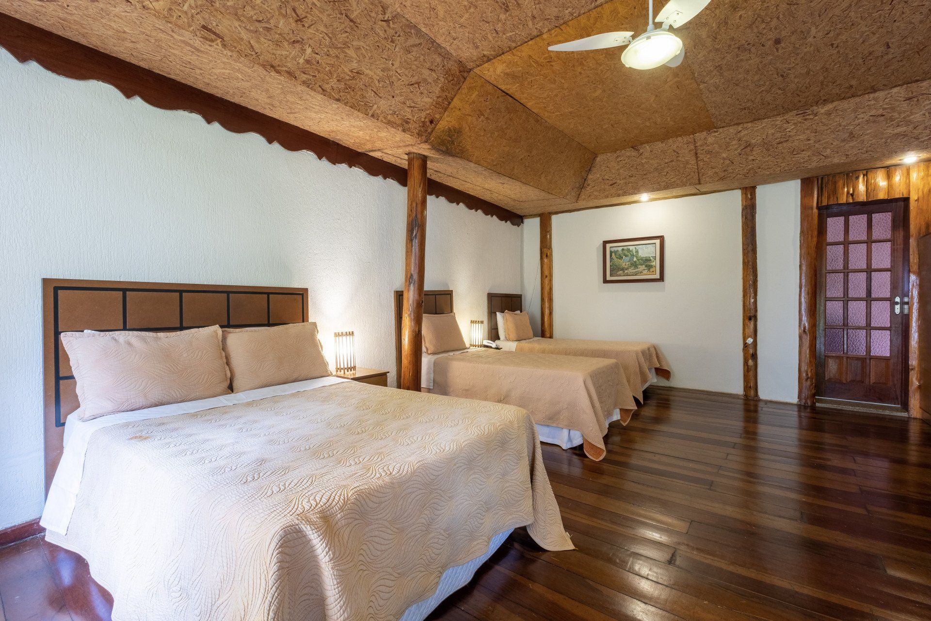 Quarto de hotel com duas camas, piso e teto de madeira. Paredes brancas e detalhes em marrom.