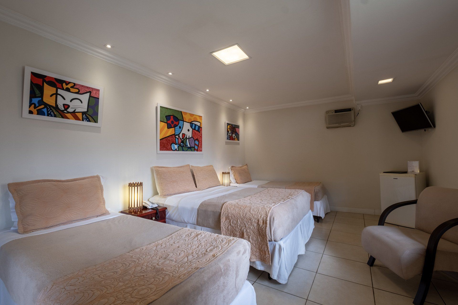 Quarto de hotel com três camas, obras de arte, frigobar e uma cadeira.
