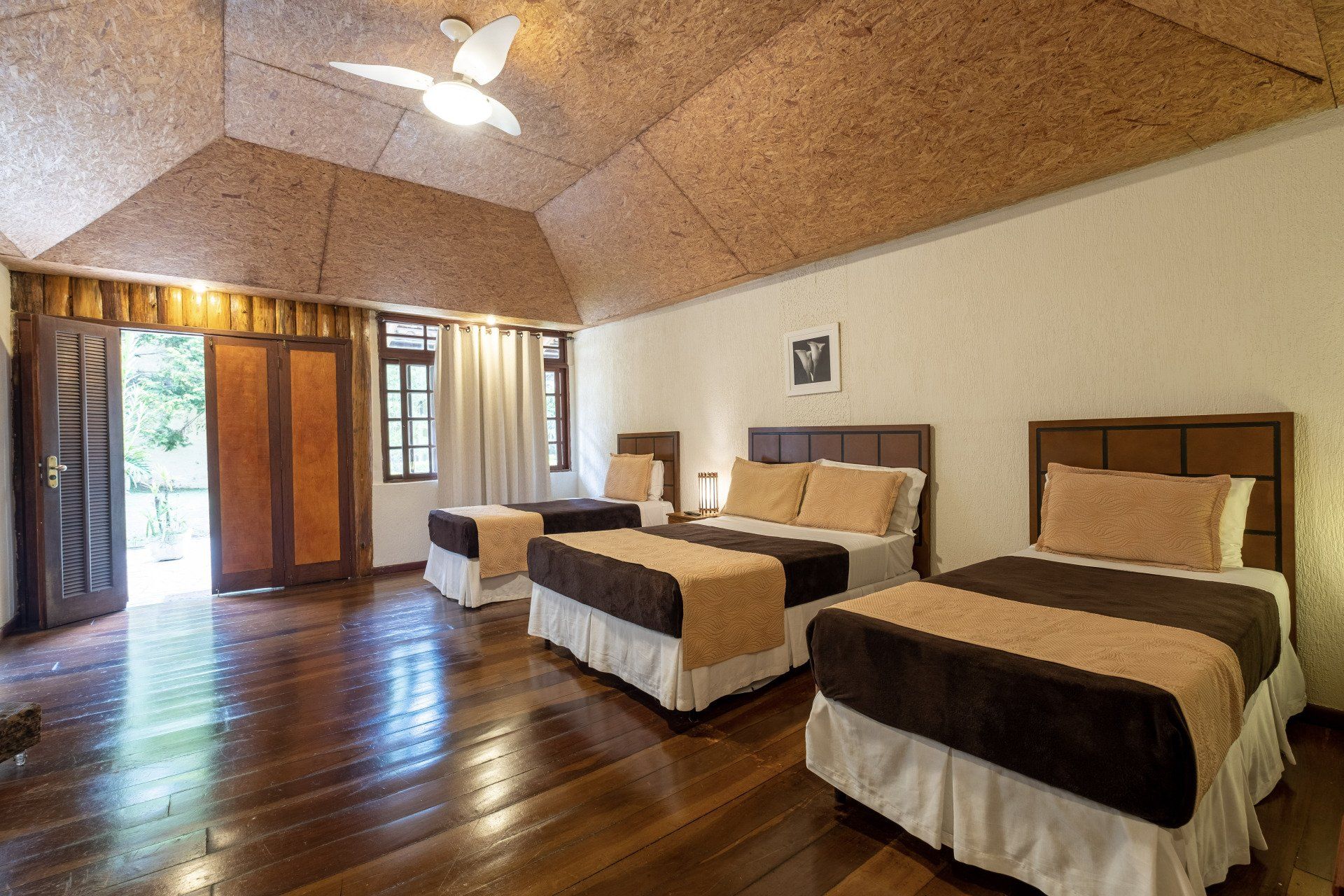 Quarto com três camas, piso e teto de madeira e portas abertas para uma varanda.