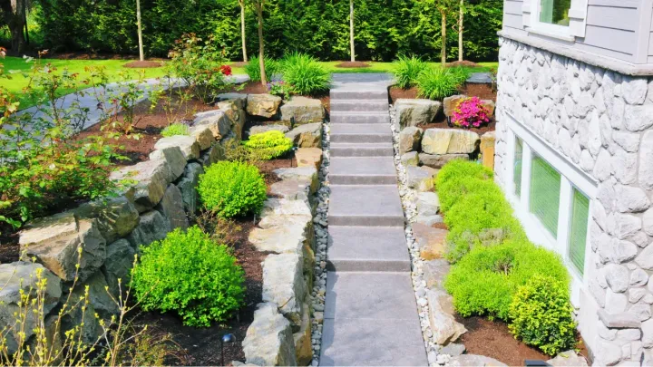 a landscaped concrete walkway with stairs in between a house and some small shrubs