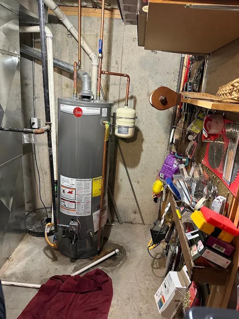 Water heater in a basement with exposed pipes and a cluttered shelf.