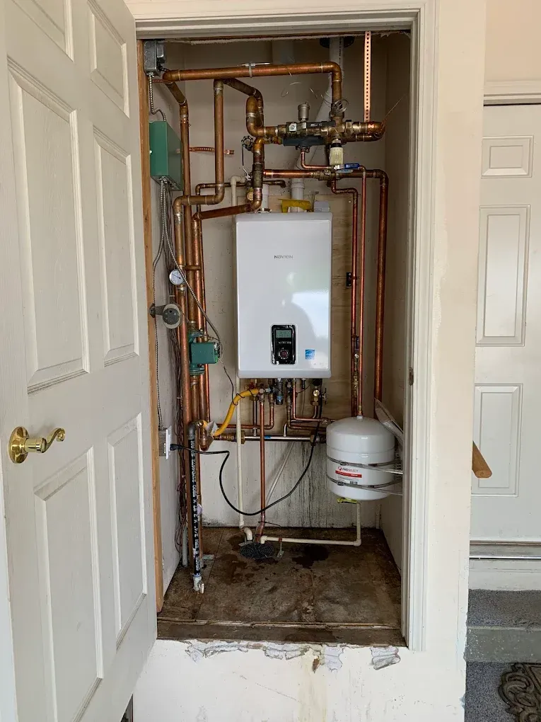 A white wall-mounted water heater with copper pipes in a small closet. Doorways visible on both sides.