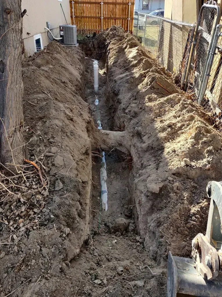 Trench dug in backyard, with white pipes installed. Soil is piled along edges. A small excavator is at the right.