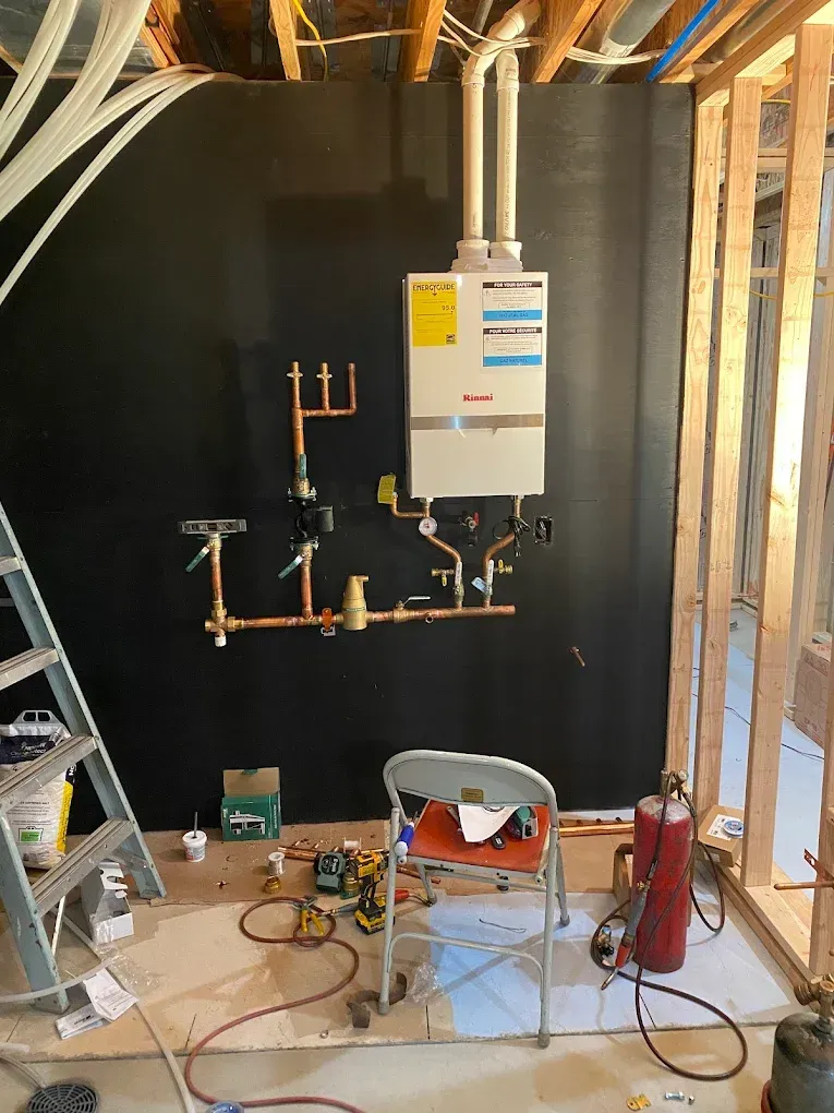 A wall-mounted water heater with copper pipes under construction in a basement. Tools and a ladder are nearby.