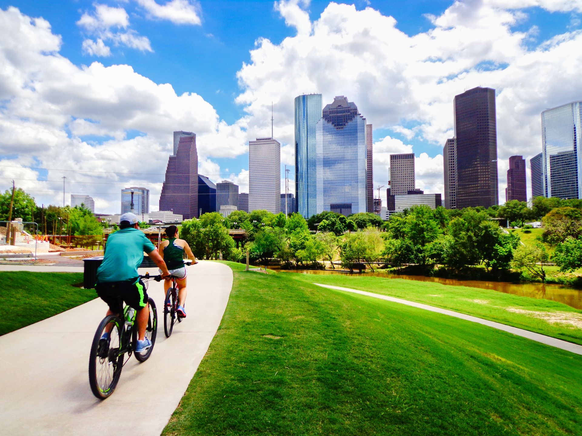 Two cyclists ride along a park path with a city skyline and bright green lawn under a cloudy blue sky.