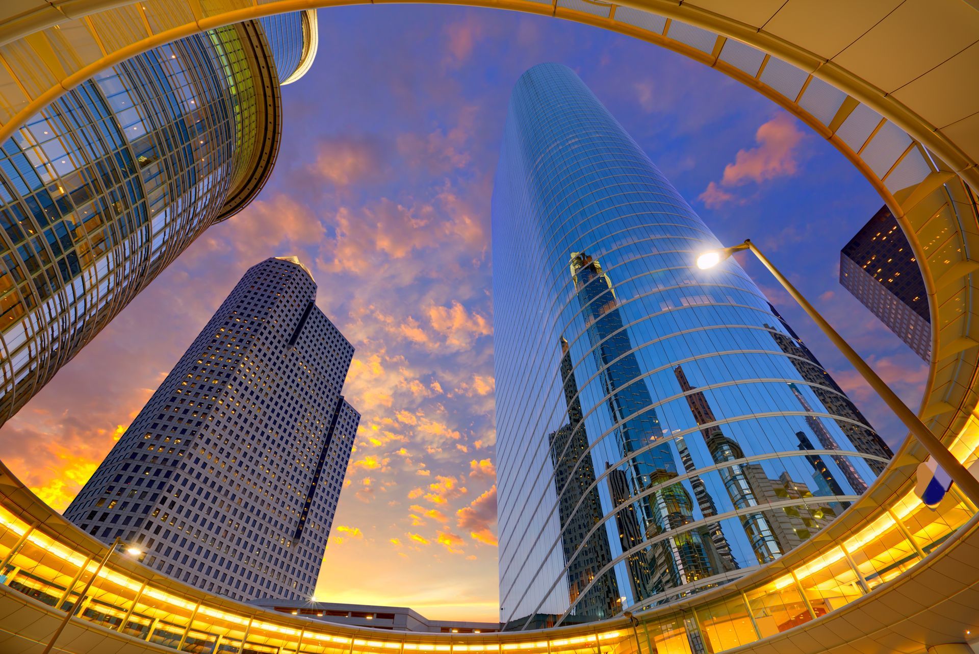 Skyscrapers reflected in a curved glass atrium at sunset, with a dramatic orange and blue sky.