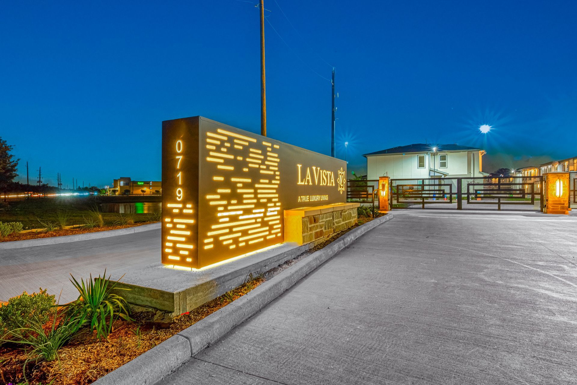 Illuminated modern entrance sign and gate at night on a paved driveway with a building in the background