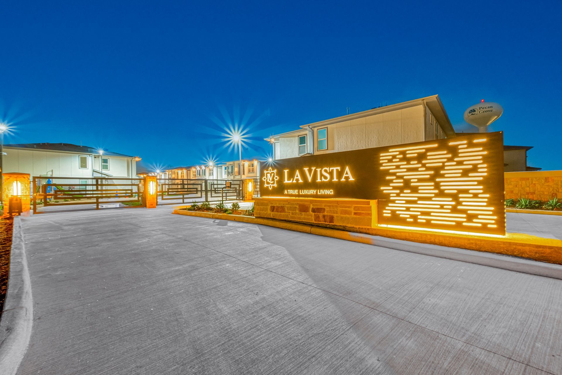 La Vista illuminated entrance sign at dusk with a wide paved driveway and buildings in the background