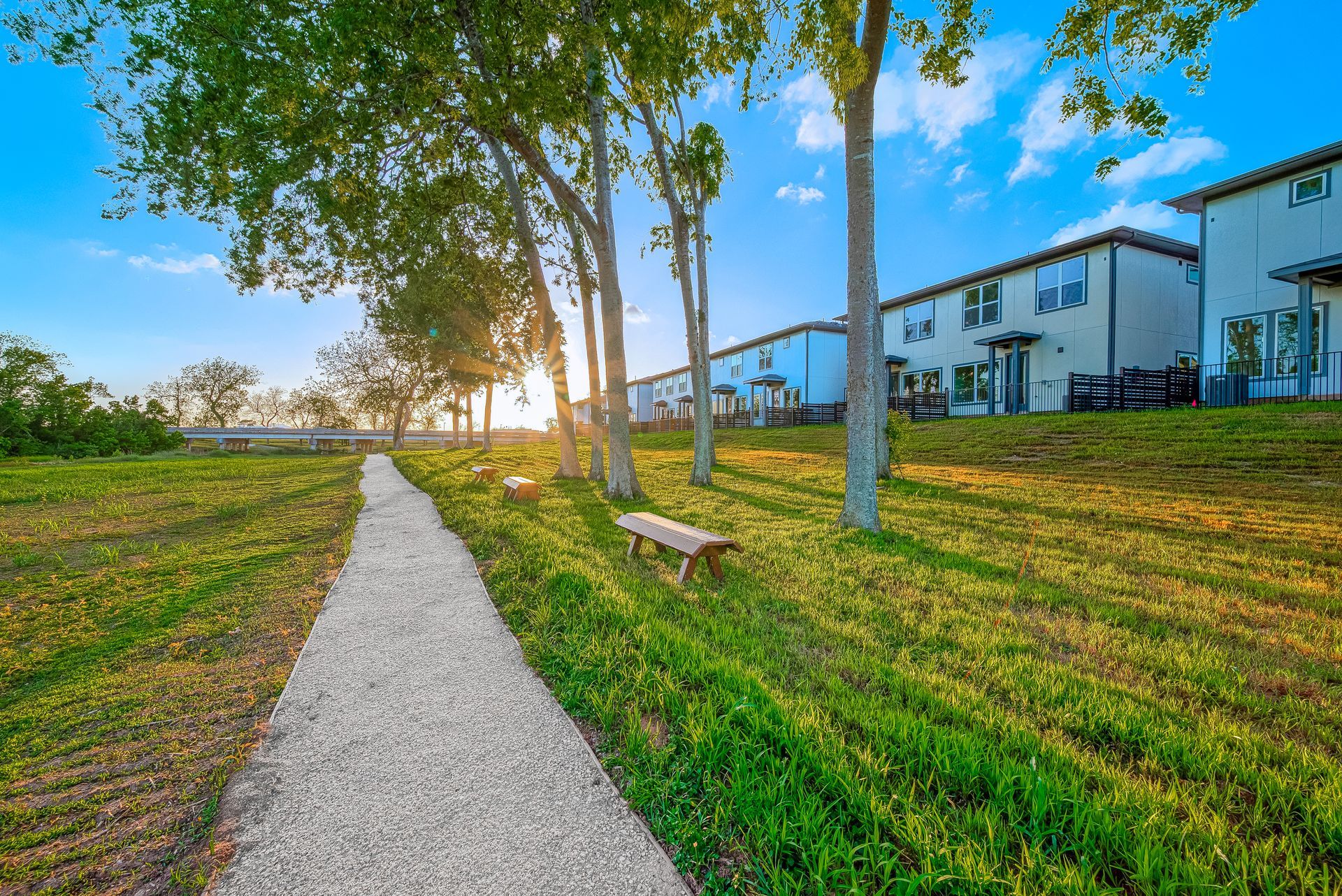 Sunlit park path beside grassy lawn, trees, bench, and modern houses at sunset