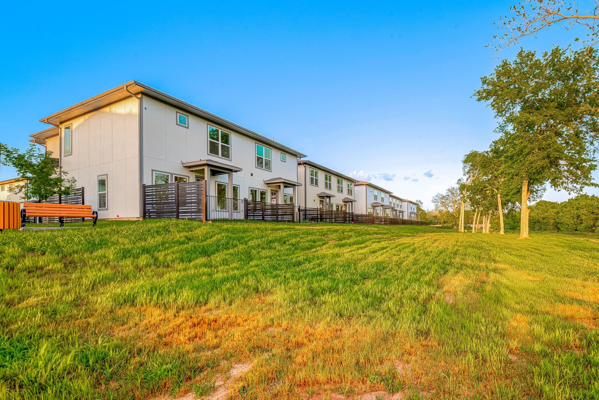 Row of beige apartment buildings beside a grassy field under a clear blue sky