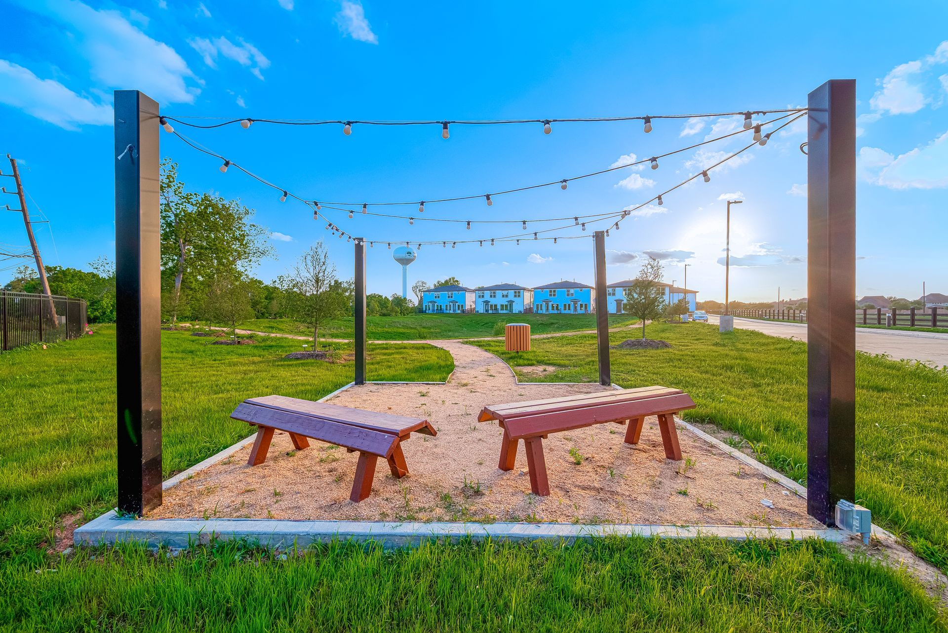 String lights over two benches in a grassy park area under a blue sky.