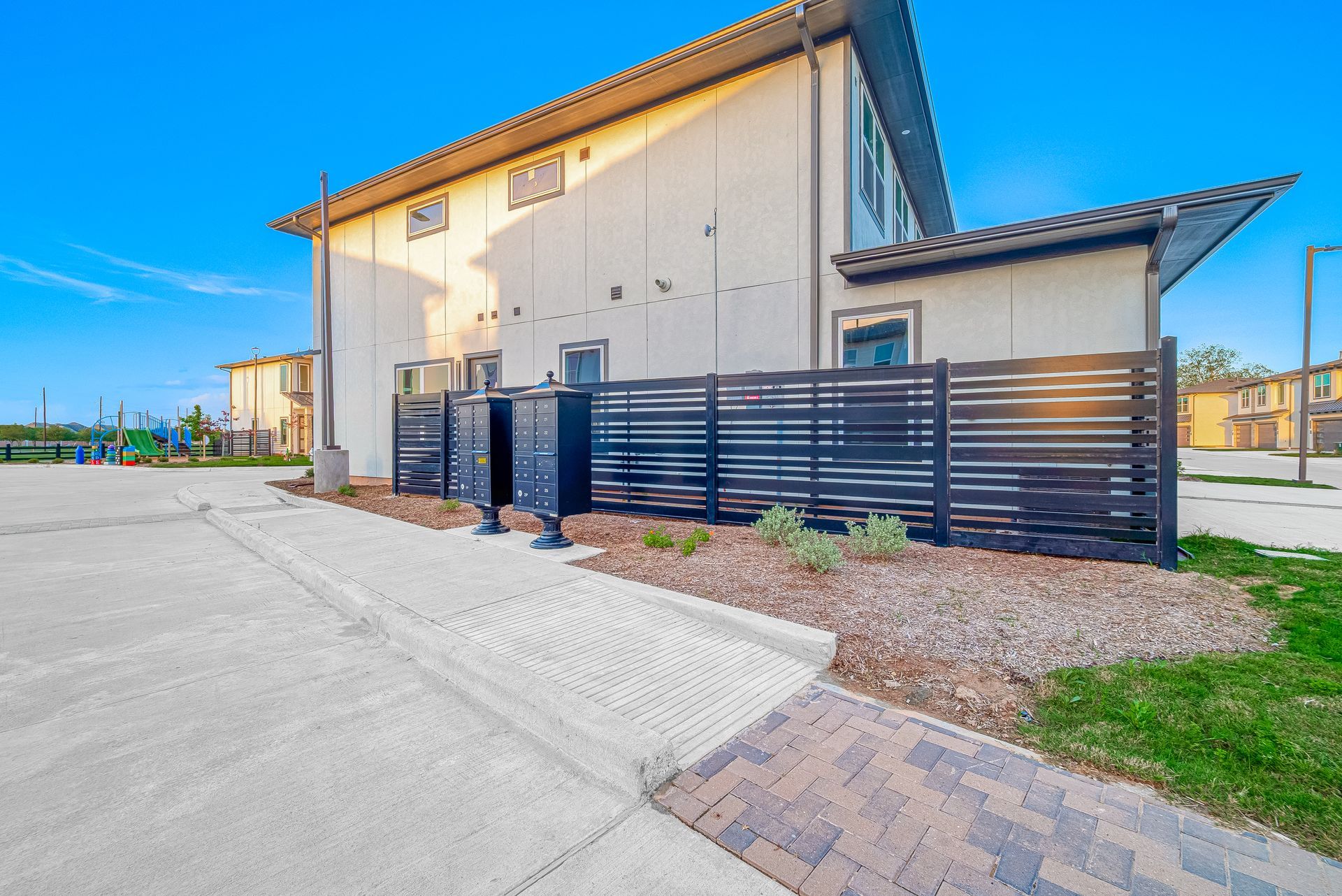 Modern beige building with black fence, paved walkway, and grassy area under a clear blue sky