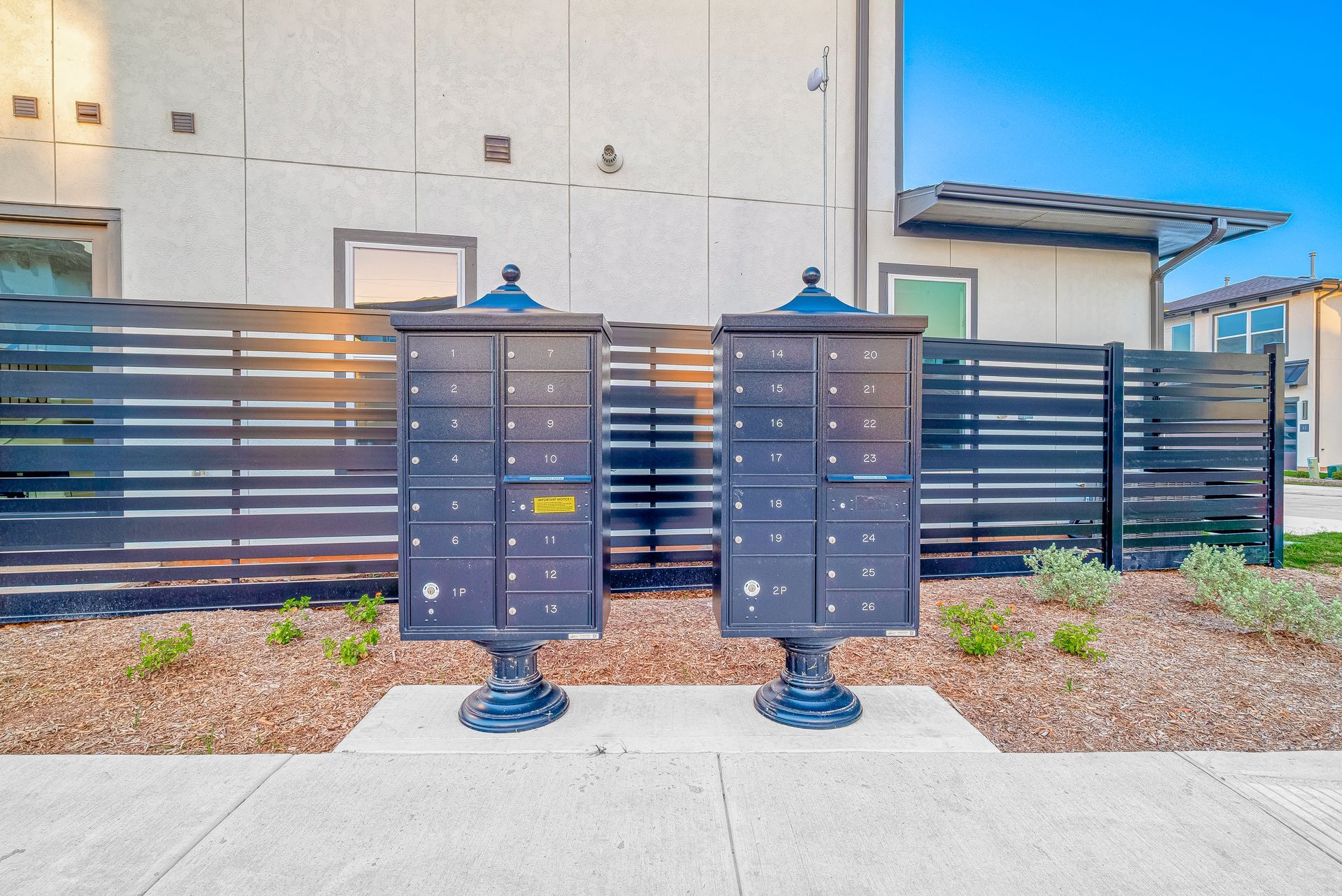 Two black mailboxes on posts in front of a modern building and dark fence, at sunset.