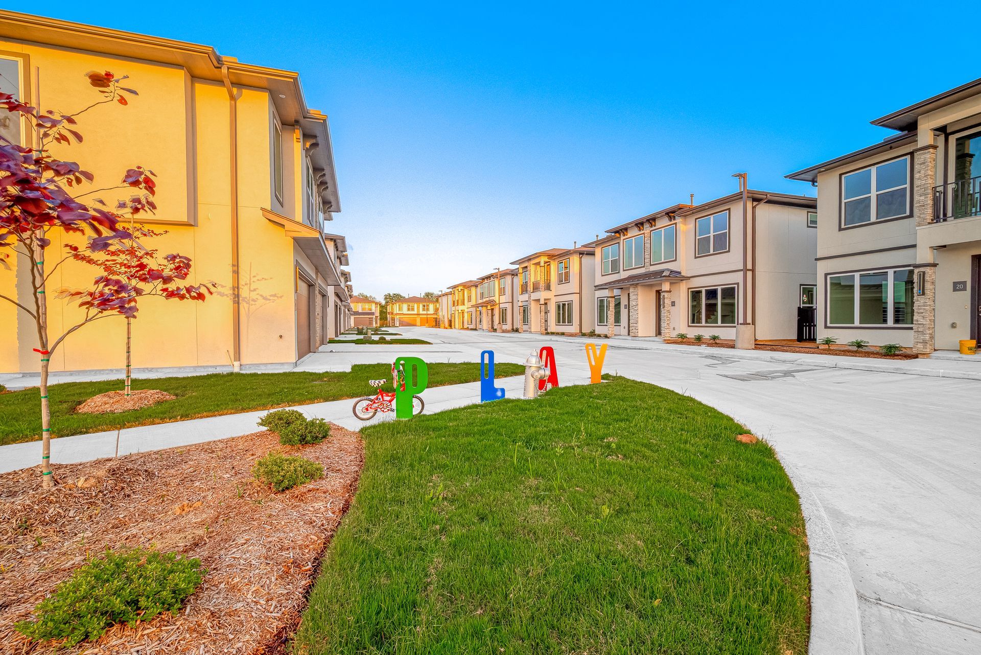 Suburban street with modern townhomes, curved sidewalk, and colorful utility boxes beside a green lawn