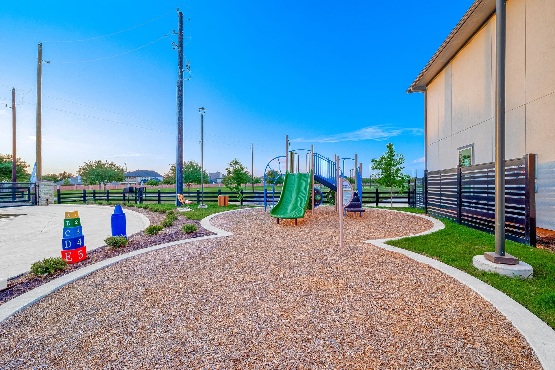 Playground with green slide, climbing structure, and pebble ground beside a building and fence