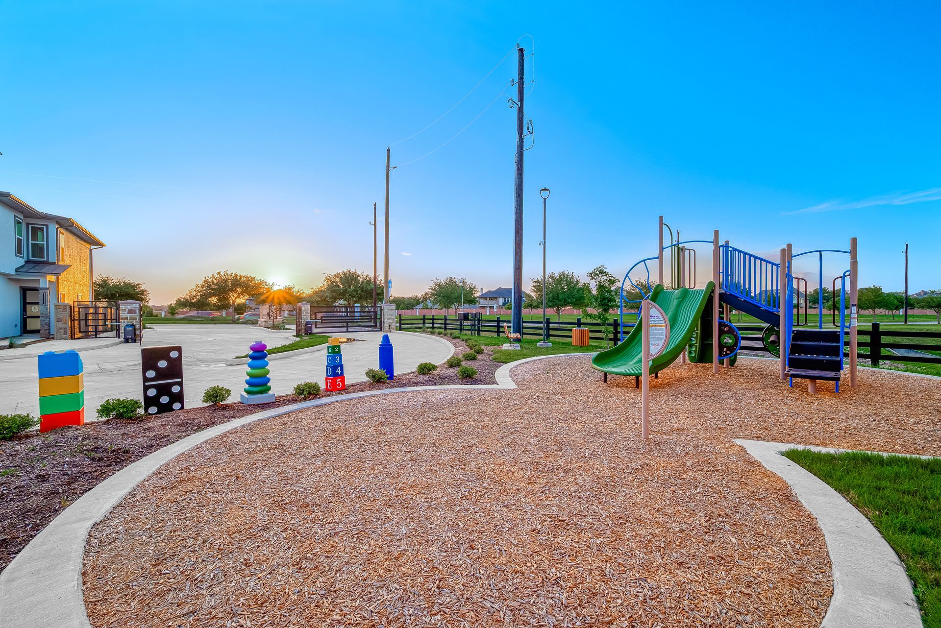 Playground with colorful climbing structures, gravel play area, and houses at sunset