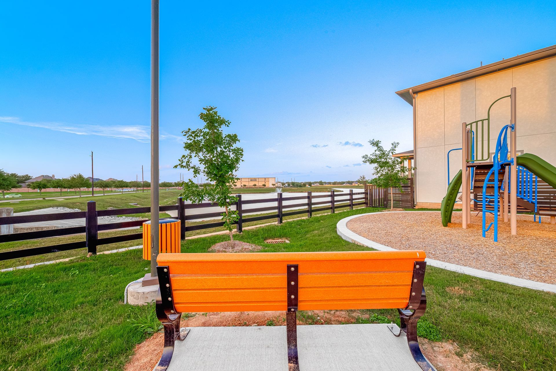 Orange bench by a grassy playground beside a beige building and blue playground equipment