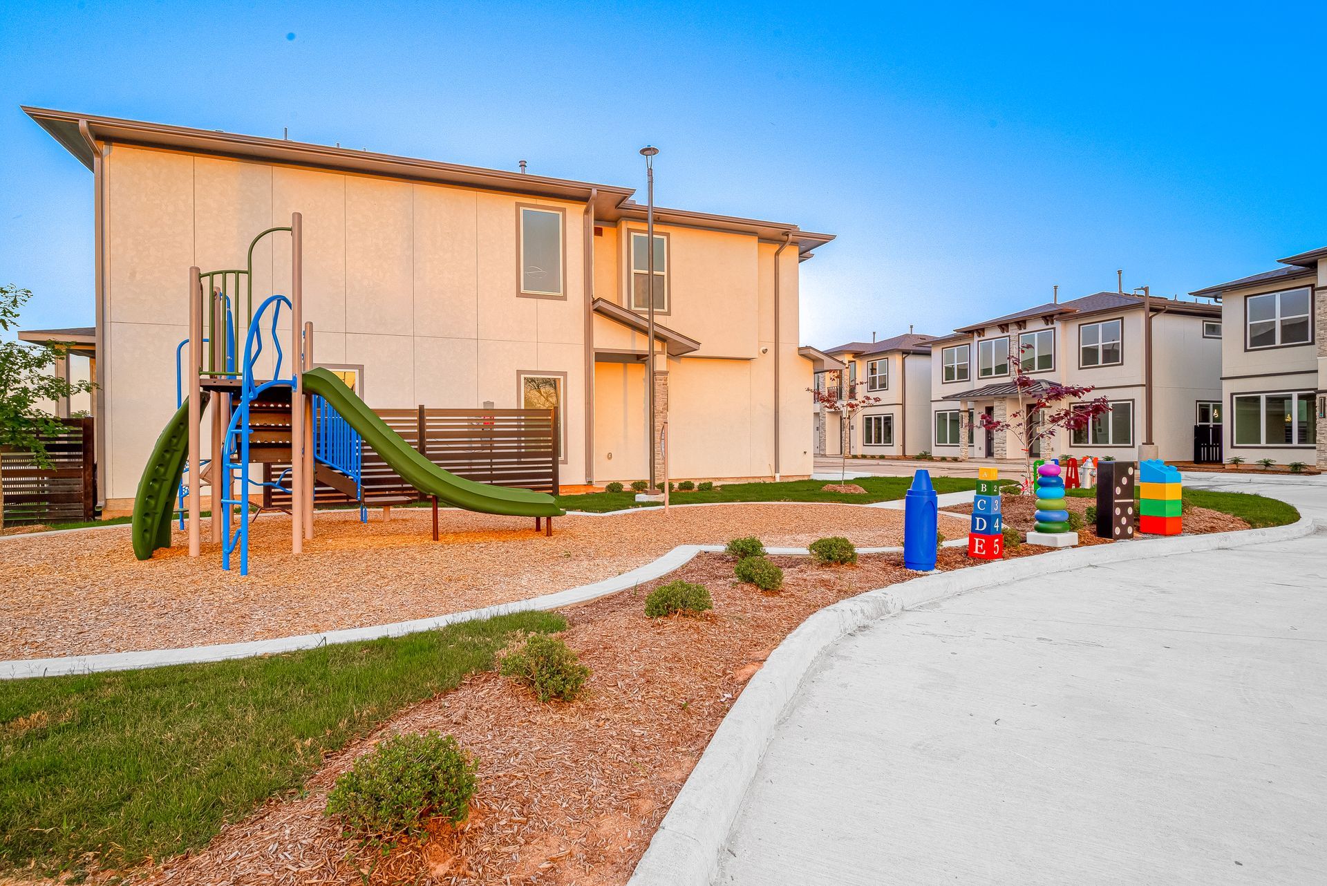 Apartment courtyard with a small playground, blue-and-green slide, and colorful play equipment on gravel and grass.