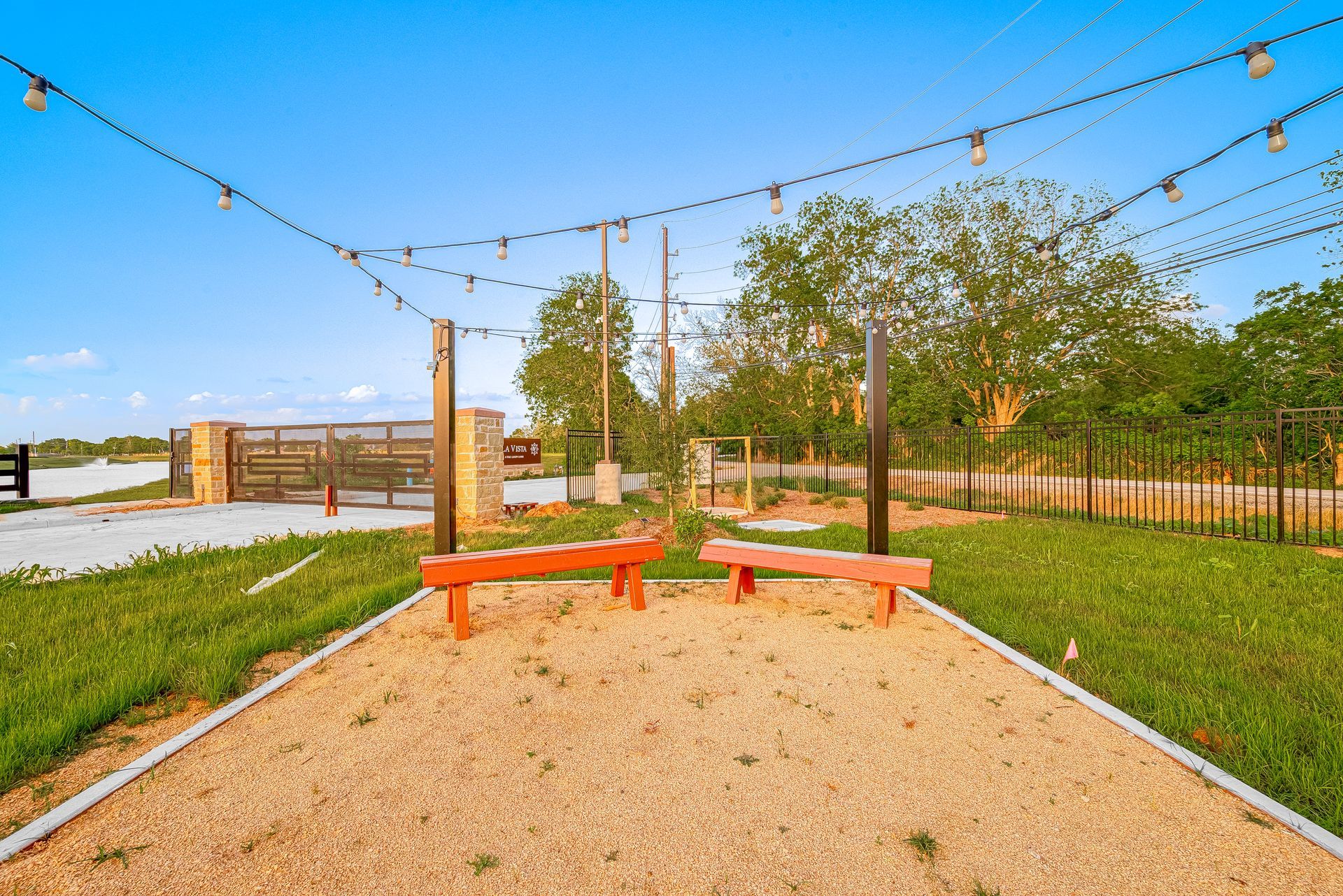 Outdoor gravel play area with orange benches, string lights, and grassy fencing under a blue sky