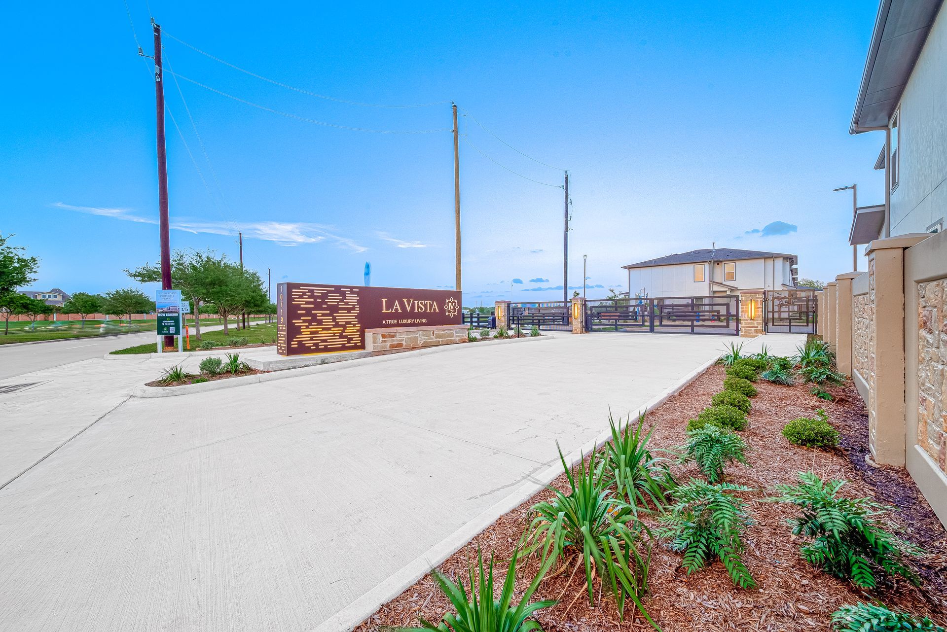 New construction site with gravel drive, wooden sign, utility poles, and landscaped planting beds under a blue sky