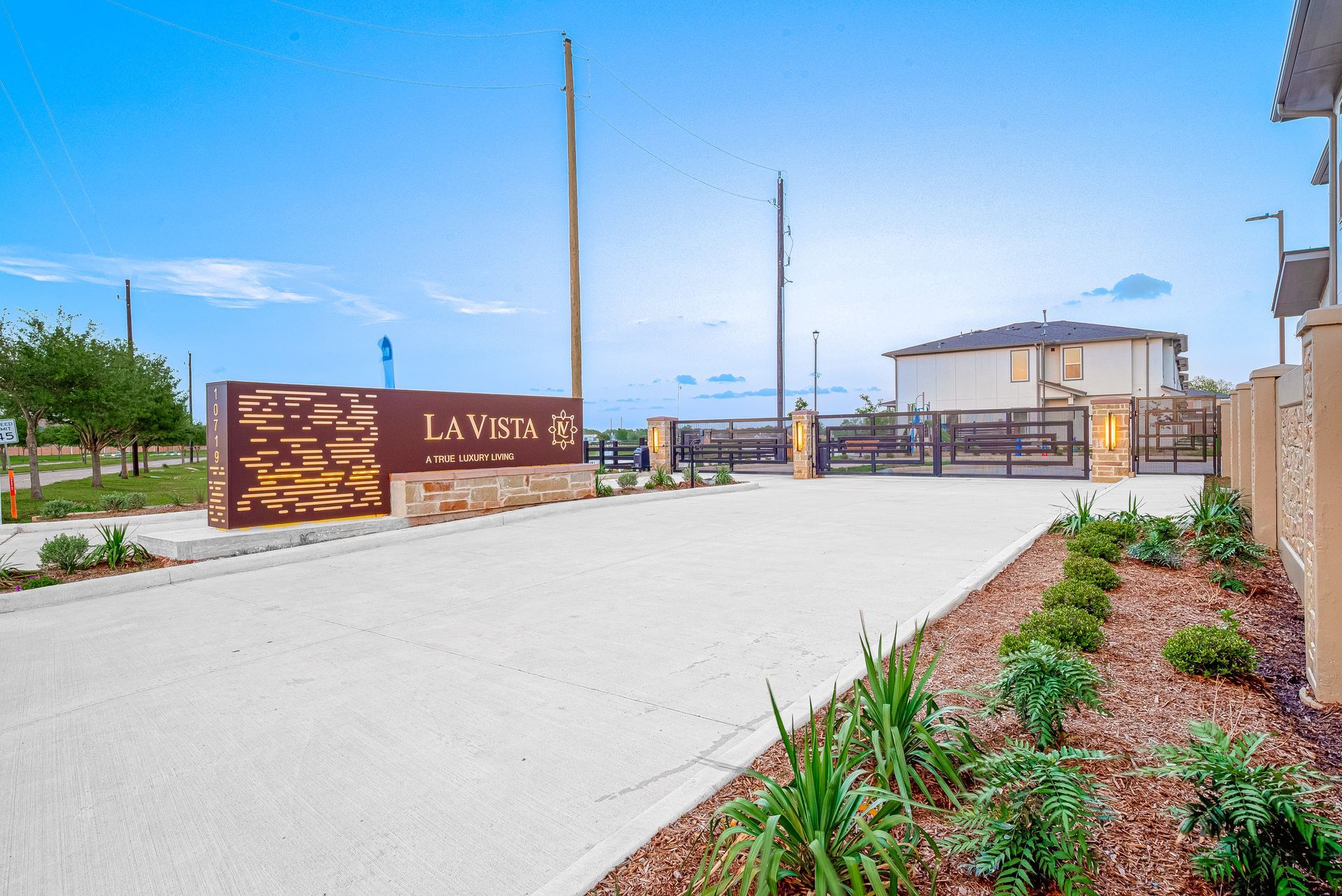 Modern gated entrance with tan walls, landscaped beds, and a clear blue sky