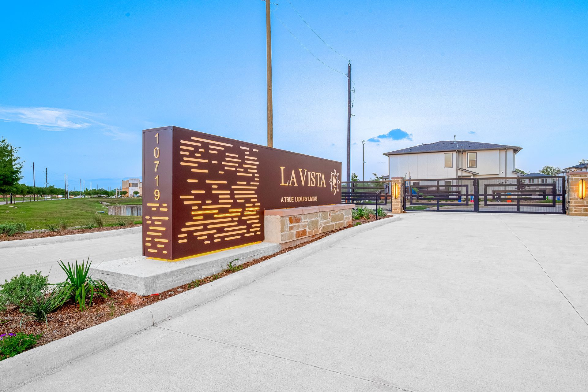 Modern gated entrance with brown signage, paved driveway, and a small building under a blue sky.
