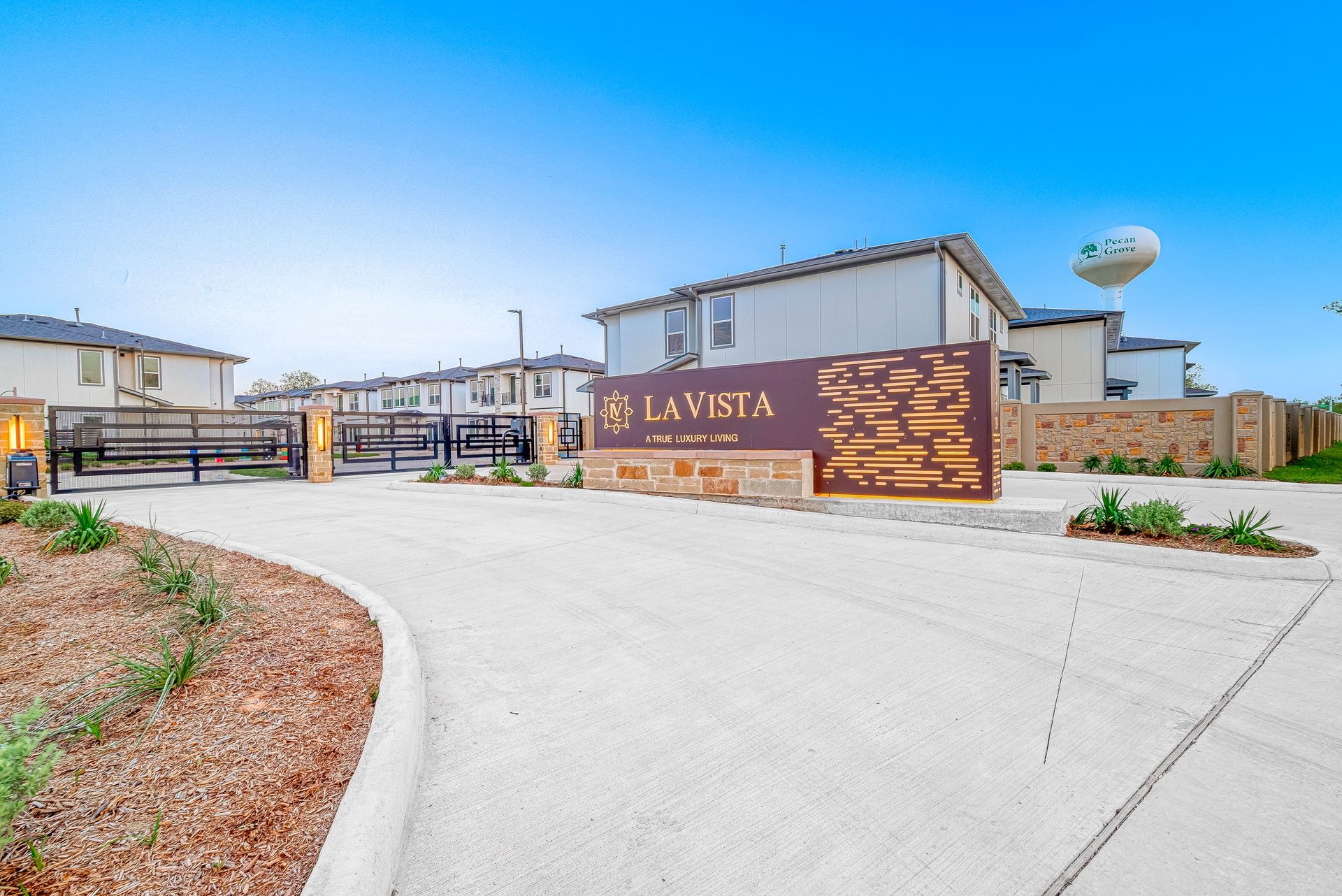 Entrance gate and sign for Lavista, with a paved road, modern homes, and a water tower under a blue sky
