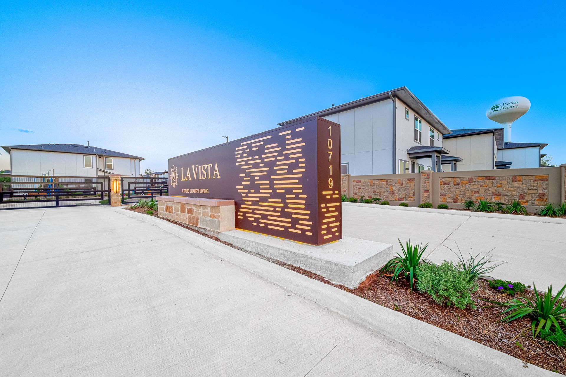 Modern apartment complex entrance with a brown sign, white walls, and landscaped plants under a blue sky