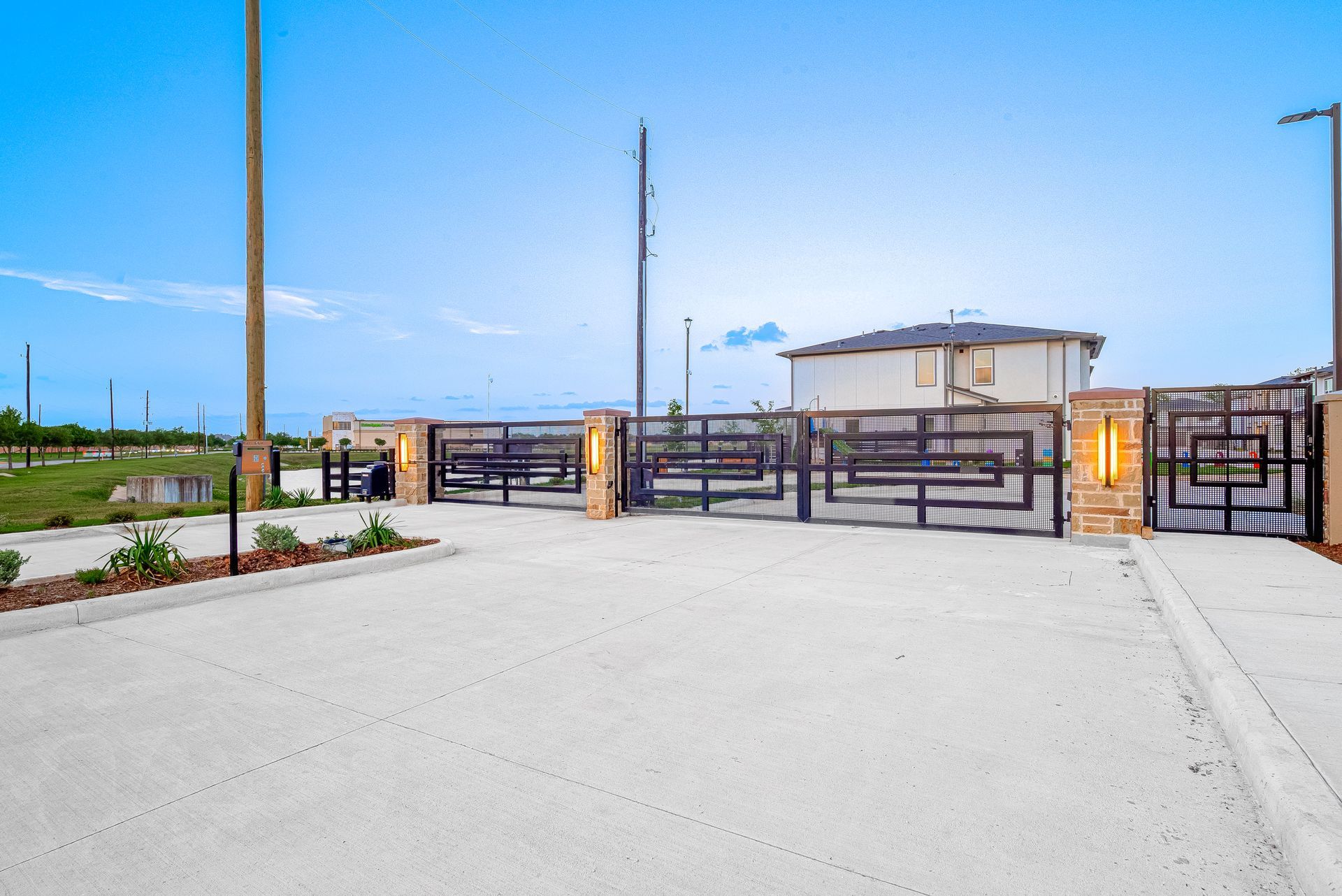 Modern gated entrance with black metal gates, stone pillars, and a wide paved driveway under a clear blue sky