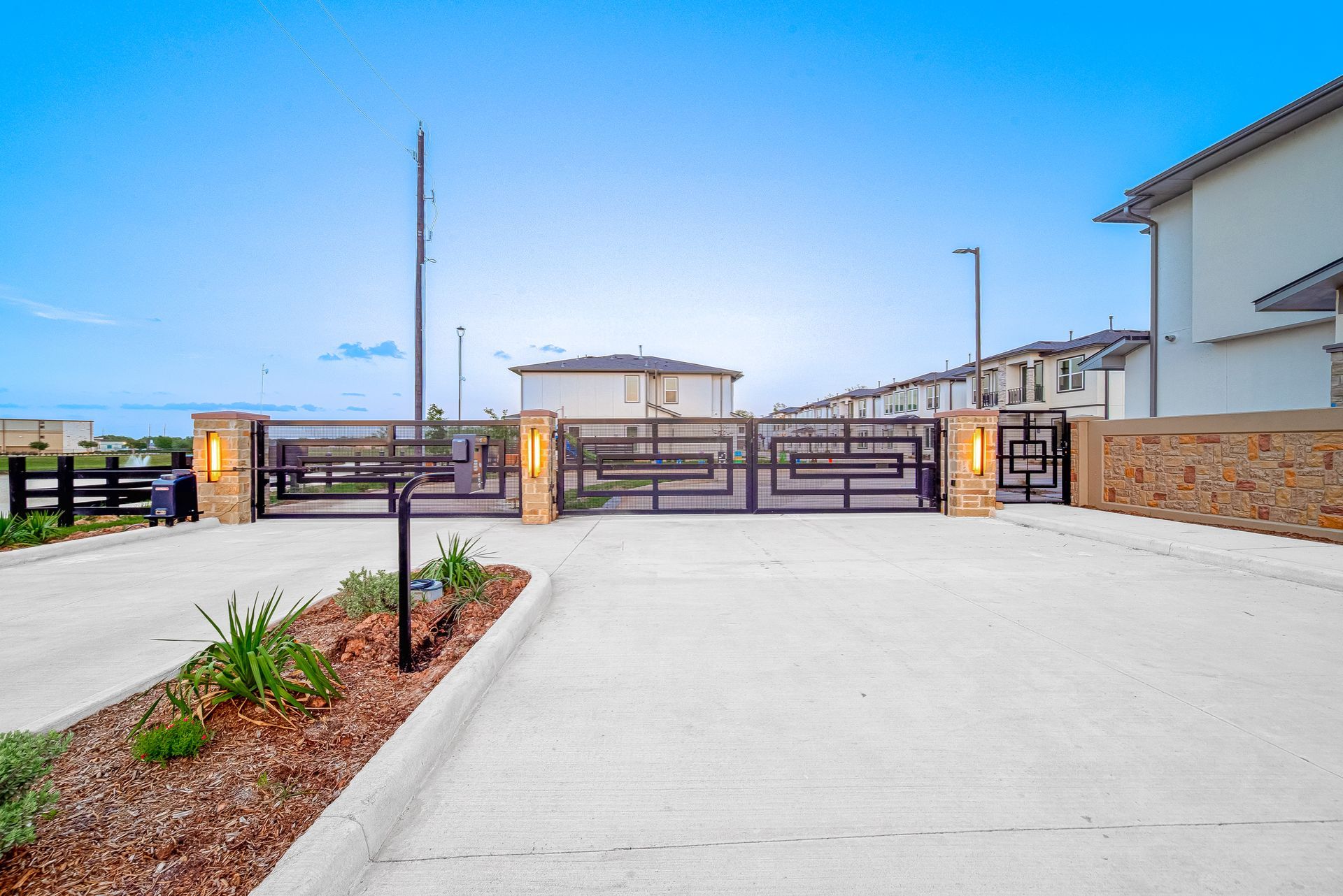 Modern driveway leading to a gated property with stone pillars and a white house under a clear sky